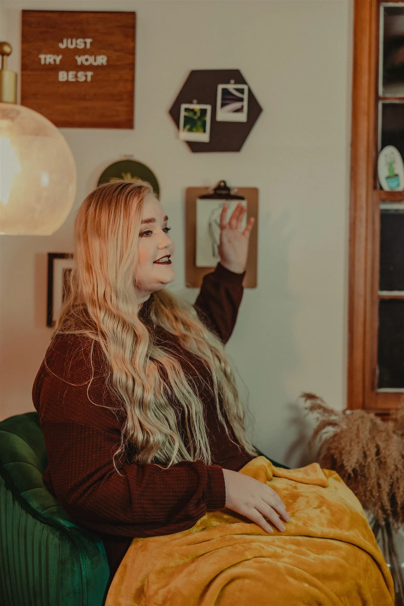 A woman with long wavy blonde hair, wearing a dark long-sleeve top and dark lipstick, sitting on a green velvet chair with a mustard-colored blanket over her lap, smiling and gesturing during a trauma informed photography session in Columbia, MO.