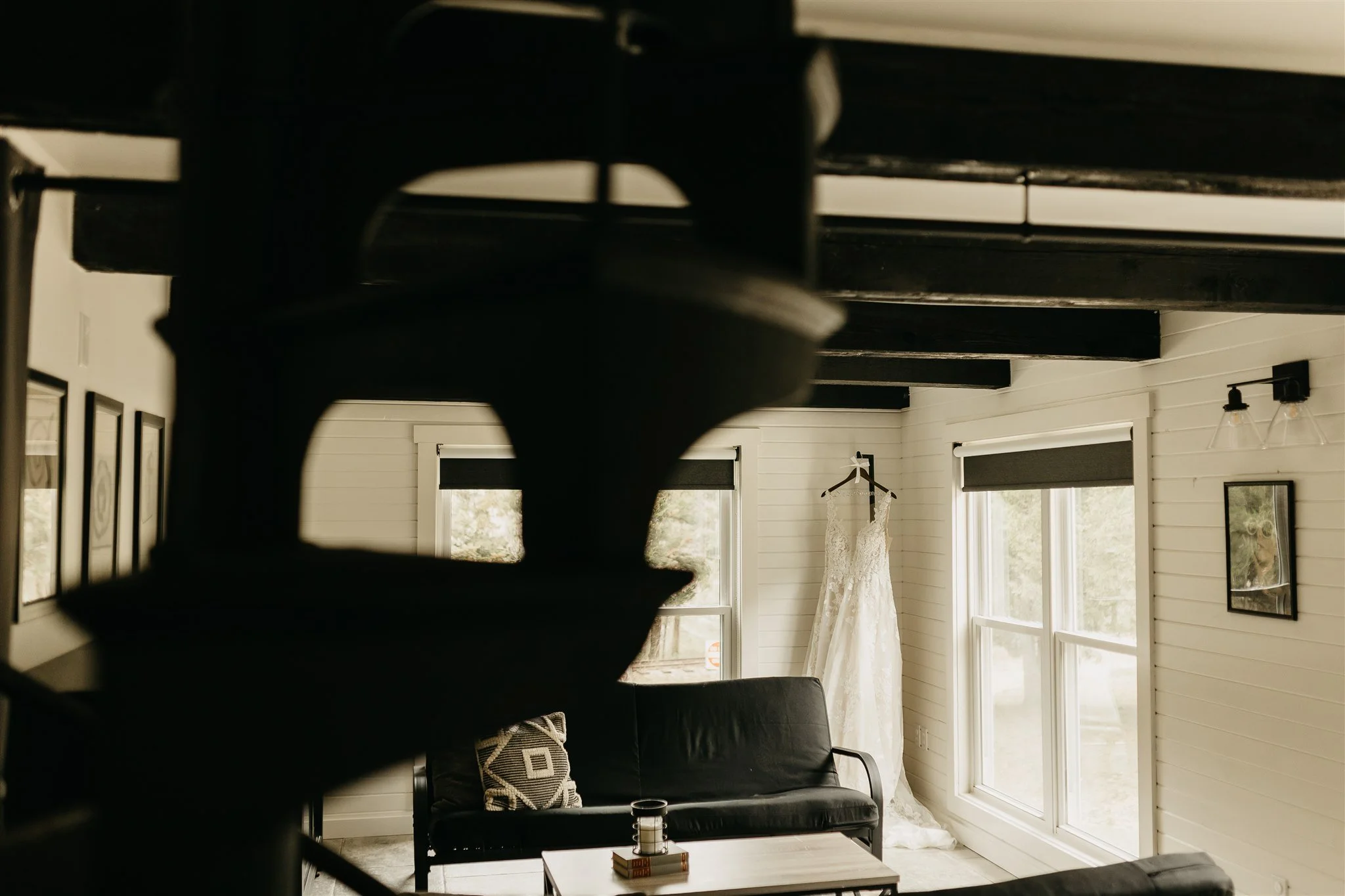 A white wedding dress hanging on a hanger in a room with white walls and large windows, with a black sofa and a coffee table in the foreground at Rolla, MO rustic wedding venue, The Village.