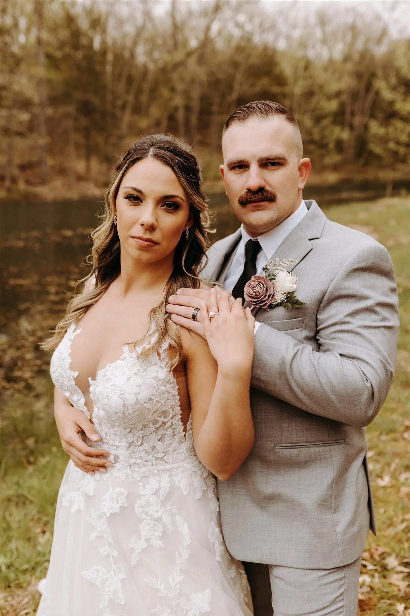 A newlywed couple in wedding attire, standing outdoors near a body of water with trees in the background at Rolla, MO rustic wedding venue, The Village. Photos by Columbia, MO Photographer Liv Strange Photography.