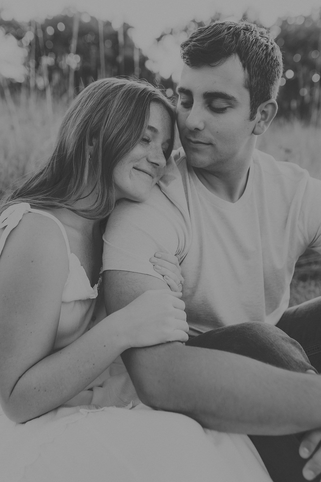 A black and white photo of a young couple sitting close together outdoors, smiling and appearing happy.