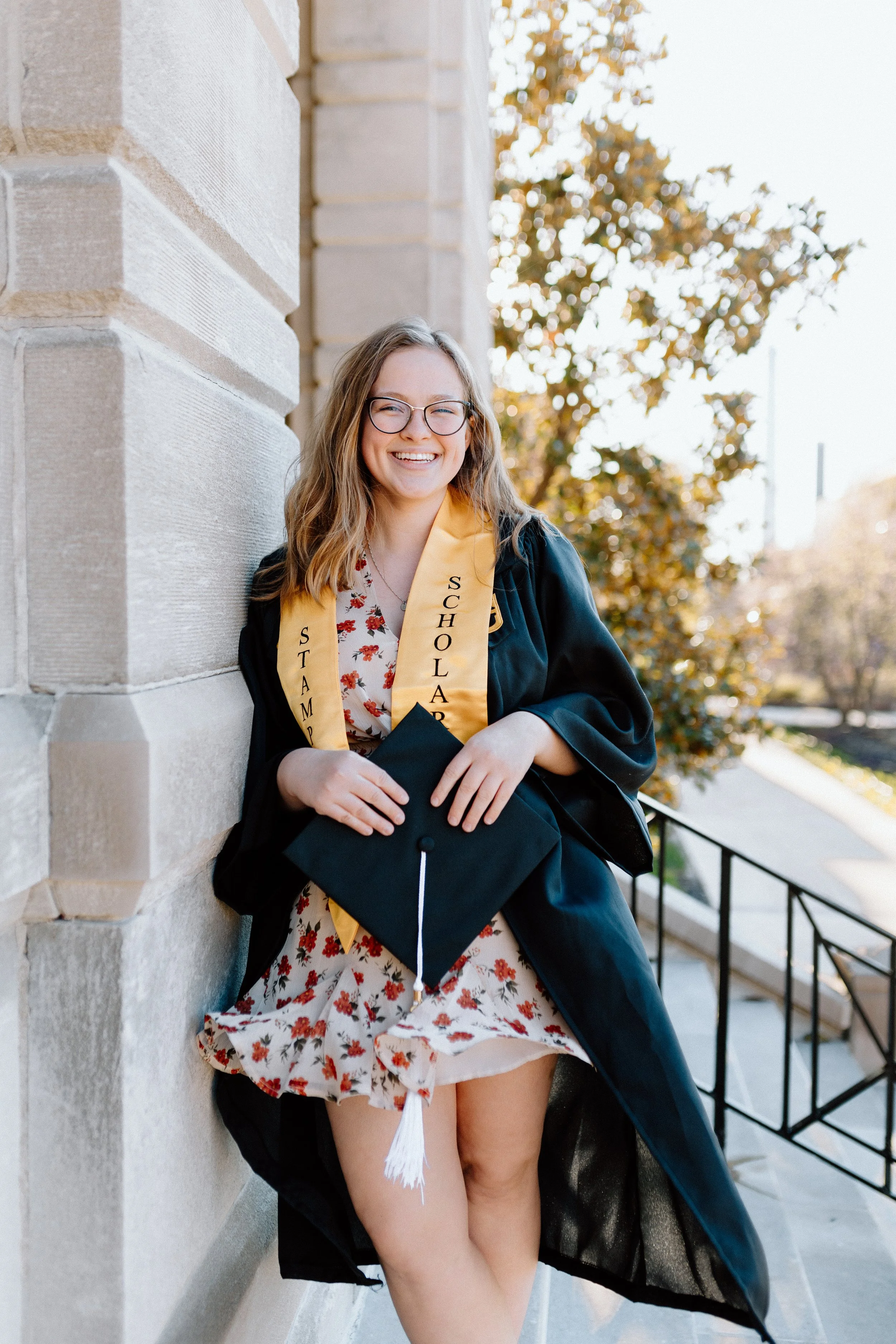 A young woman in graduation attire, wearing a black gown and a yellow stole, smiling and holding a black mortarboard cap, standing against a stone building with trees and clear sky in the background.
