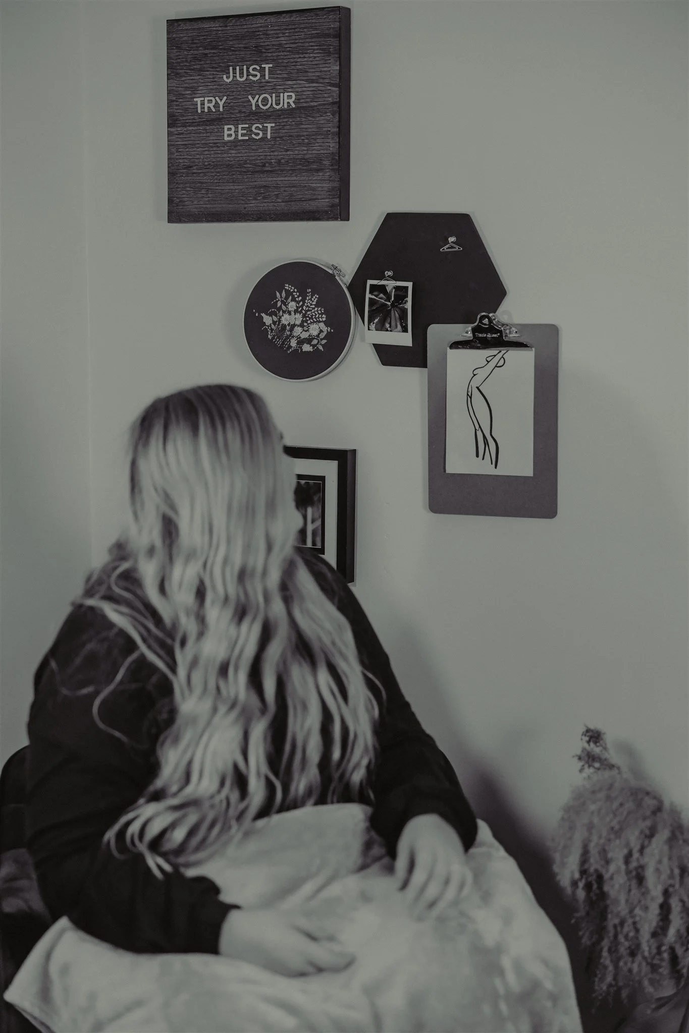 A woman with long wavy hair sitting on a bed in front of a wall decorated with various black framed items. There is a gray blanket on her lap, and a fluffy dog sitting on the floor nearby during a trauma informed photography session in Columbia, MO.