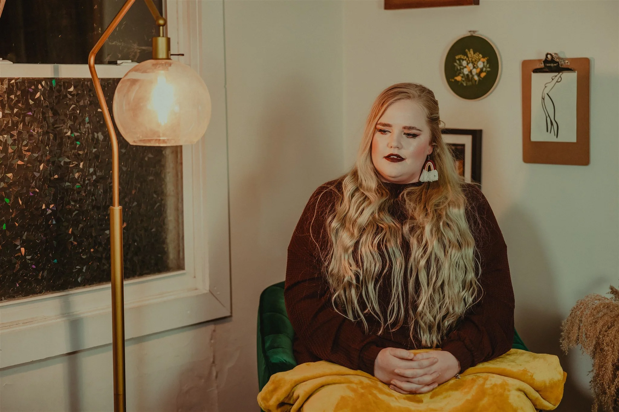 A woman with long, wavy blonde hair sits on a green chair, wearing a maroon sweater and dark lipstick, holding her hands together in her lap. There is a yellow blanket on her lap during a trauma informed photography session in Columbia, Missouri.