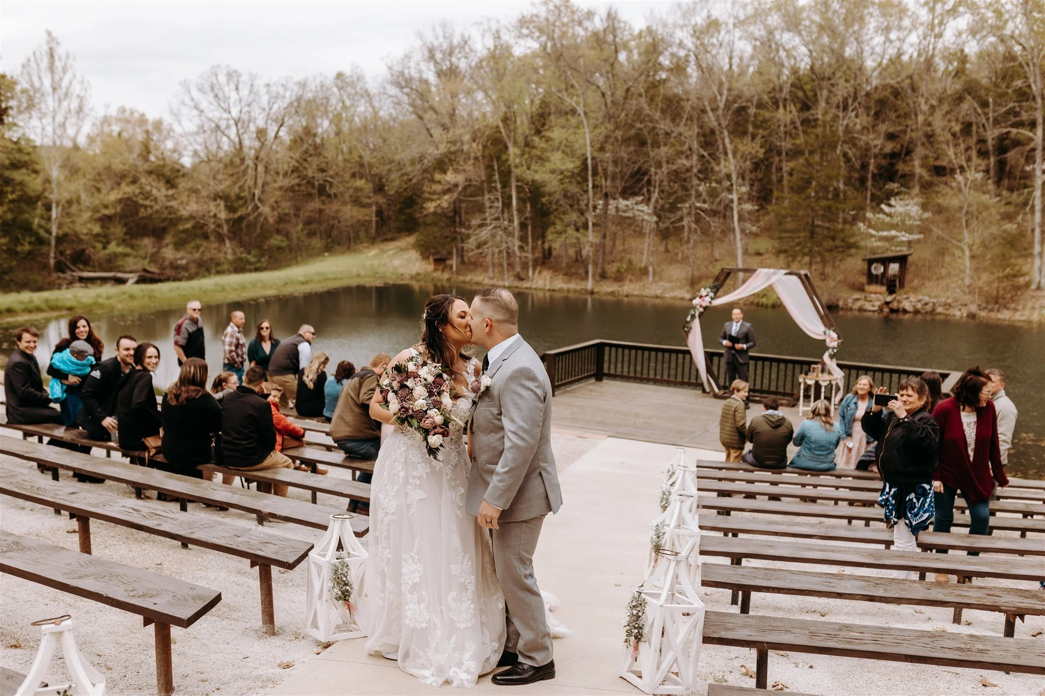A wedding ceremony taking place outdoors by a lake with a bride and groom kissing, surrounded by seated guests at Rolla, Missouri rustic wedding venue, The Village. Photos by Columbia, MO Photographer Liv Strange Photography.