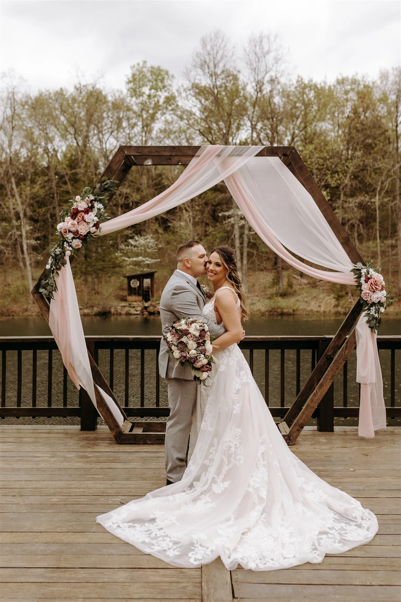 A bride and groom standing on a wooden deck by a lake, embracing and smiling at Rolla, MO rustic wedding venue, The Village. Photos by Columbia, MO Photographer Liv Strange Photography.