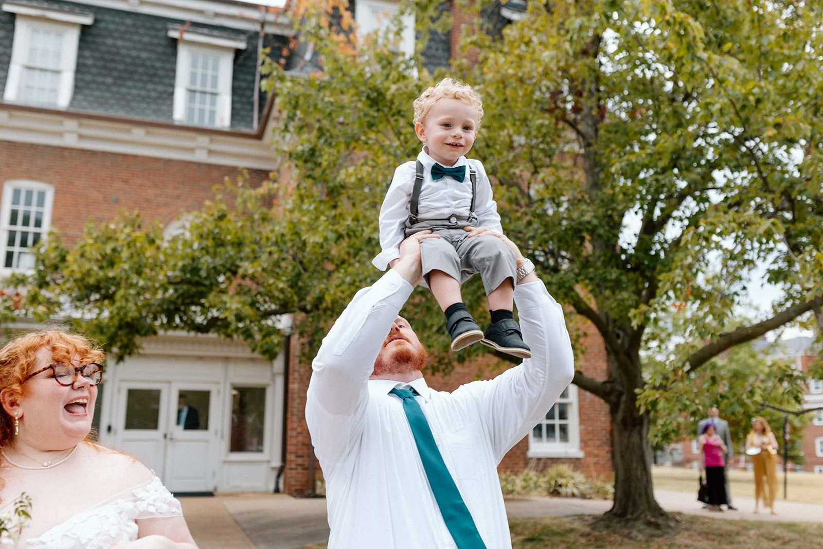 A man lifting a young boy in the air during an outdoor celebration with a woman and other people in the background, trees, and a brick building at an LGBTQ+ wedding in Columbia, MO at wedding venue, Stephens College Senior Hall.