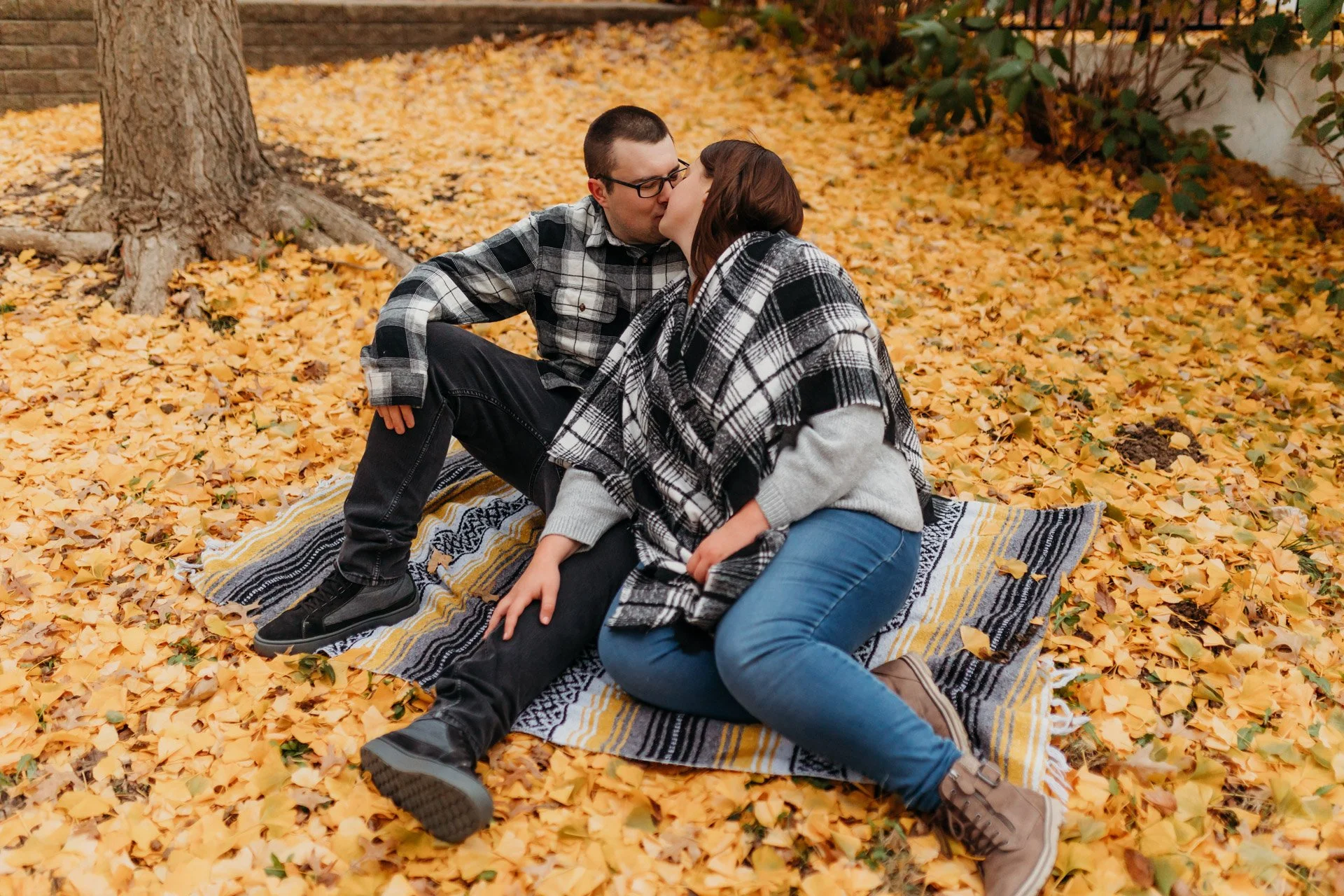Couple sharing a kiss on a blanket in a park surrounded by fallen yellow leaves in autumn at their engagement photography session at Queeny Park in STL.
