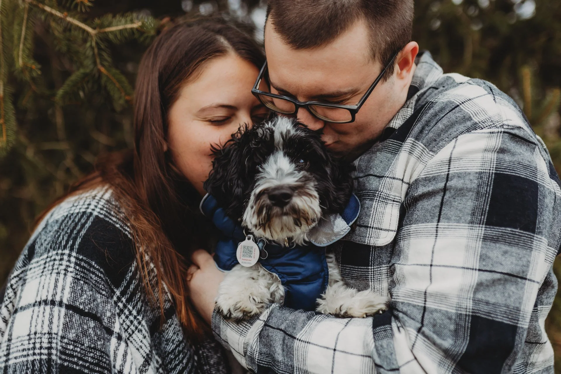 A couple holding a black and white puppy dressed in a blue jacket, embracing it affectionately outdoors with a background of green foliage at their engagement photography session at Queeny Park in STL.
