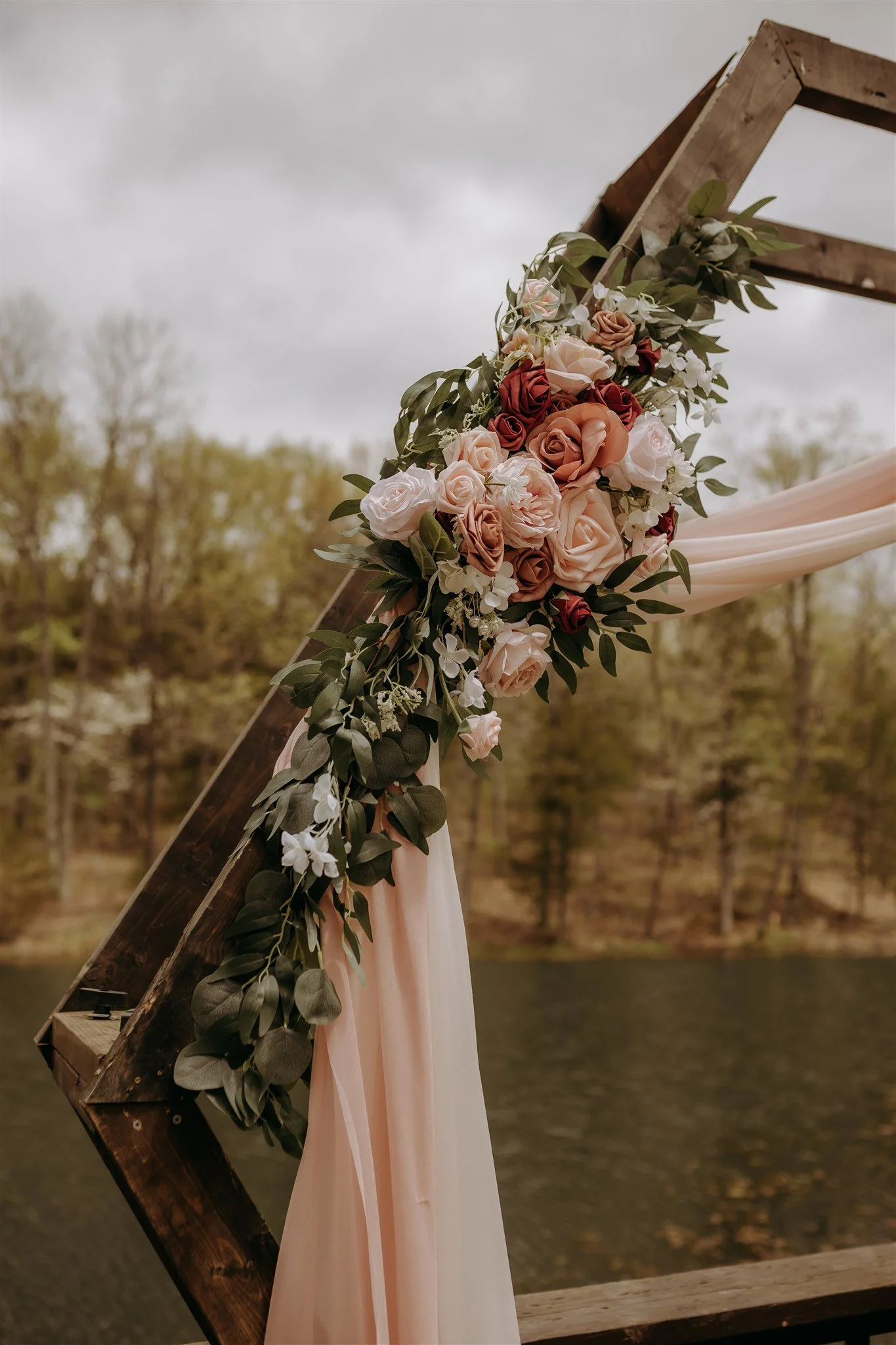Decorated wooden arch with pink, peach, and burgundy roses, green foliage, and draped fabric, outdoors near a lake with trees in the background at Rolla, MO rustic wedding venue, The Village.