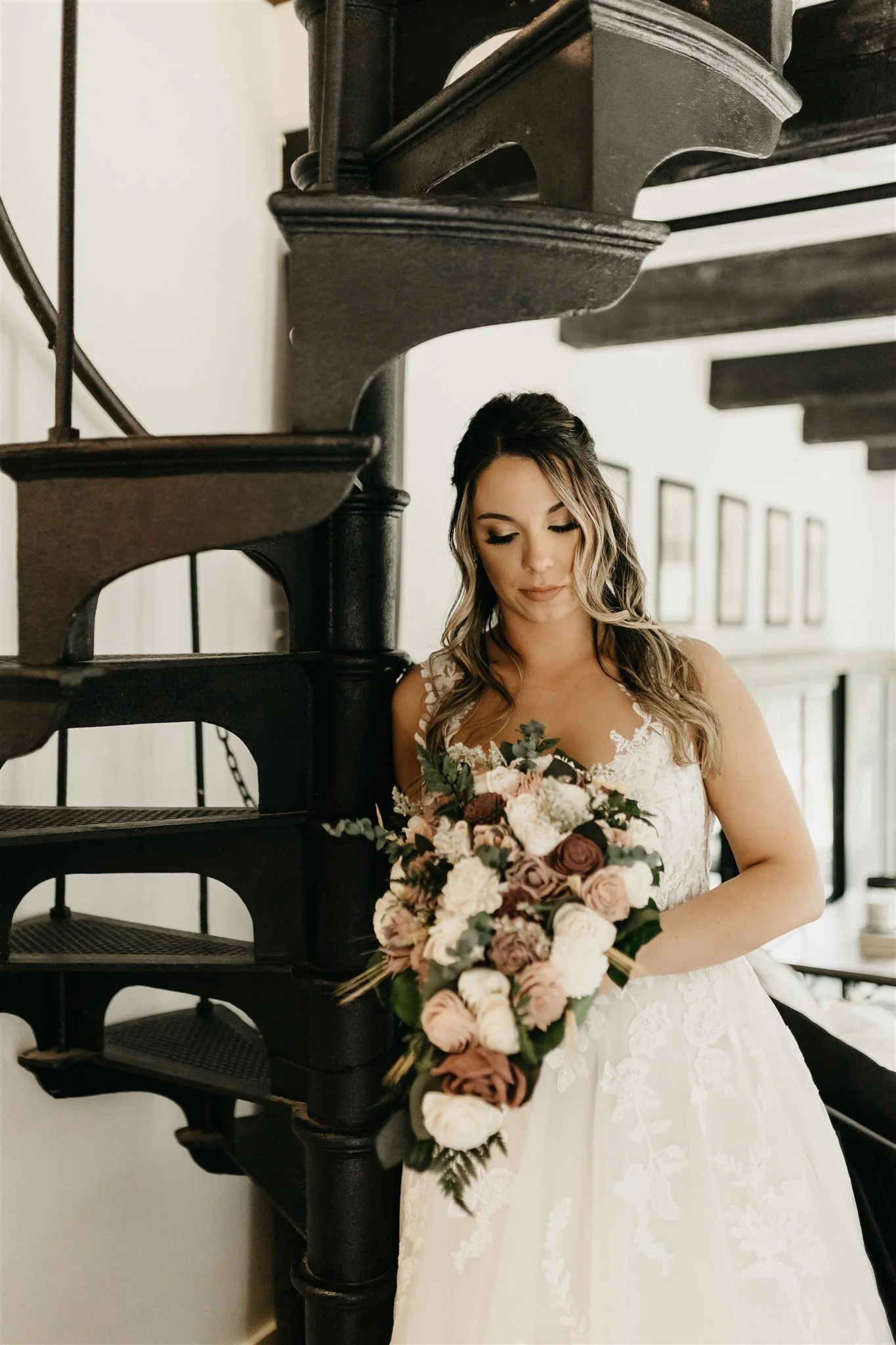 Portrait of a bride in a white wedding dress holding a bouquet, standing next to a black spiral staircase in a well-lit room at Rolla, Missouri rustic wedding venue, The Village. Photos by Columbia, MO Photographer Liv Strange Photography.