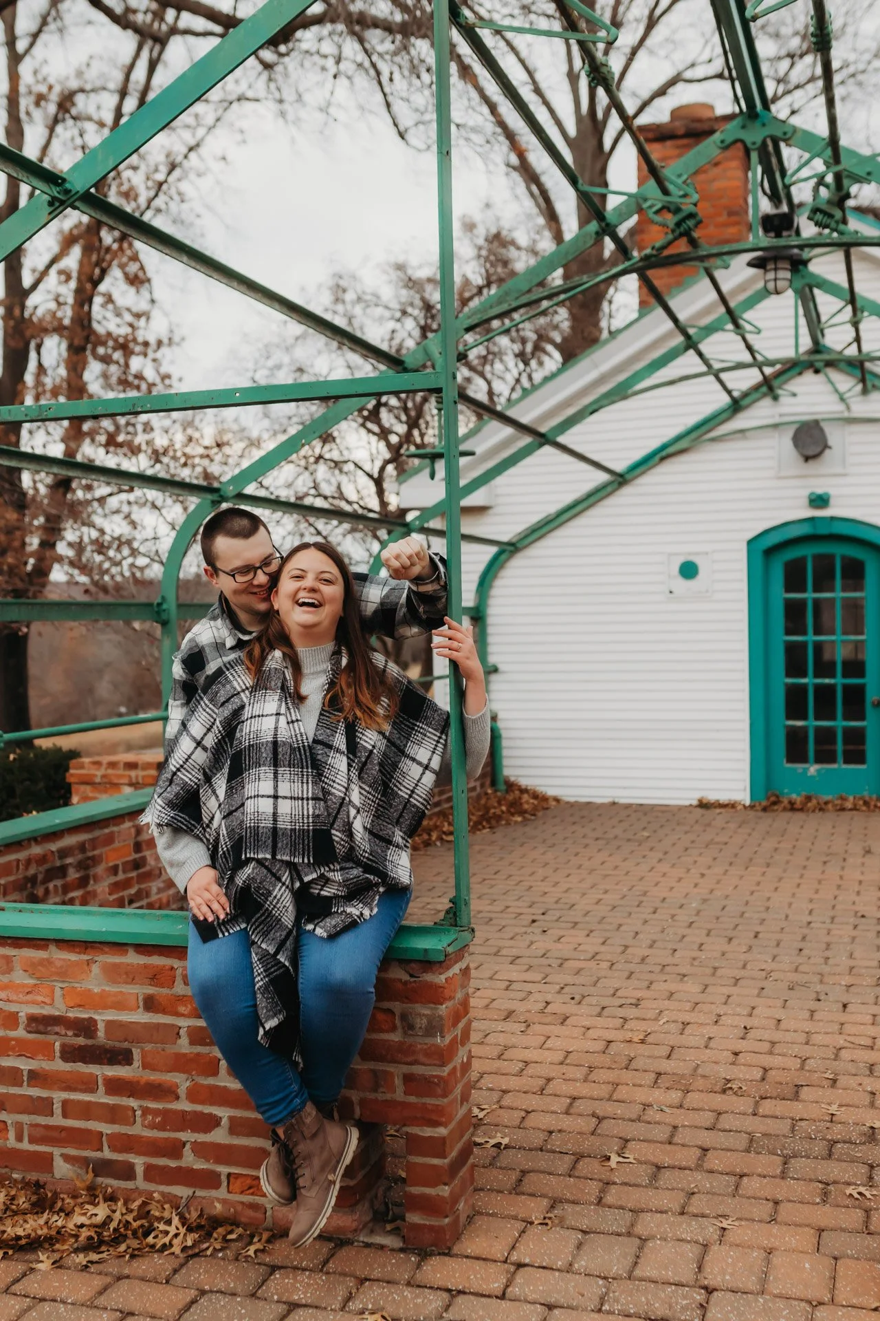 A happy couple stands outside on brick pavement, leaning on a brick corner with green metal framing above them. The woman is laughing and the man is smiling, and has his arm around her at their engagement photography session at Queeny Park in STL.