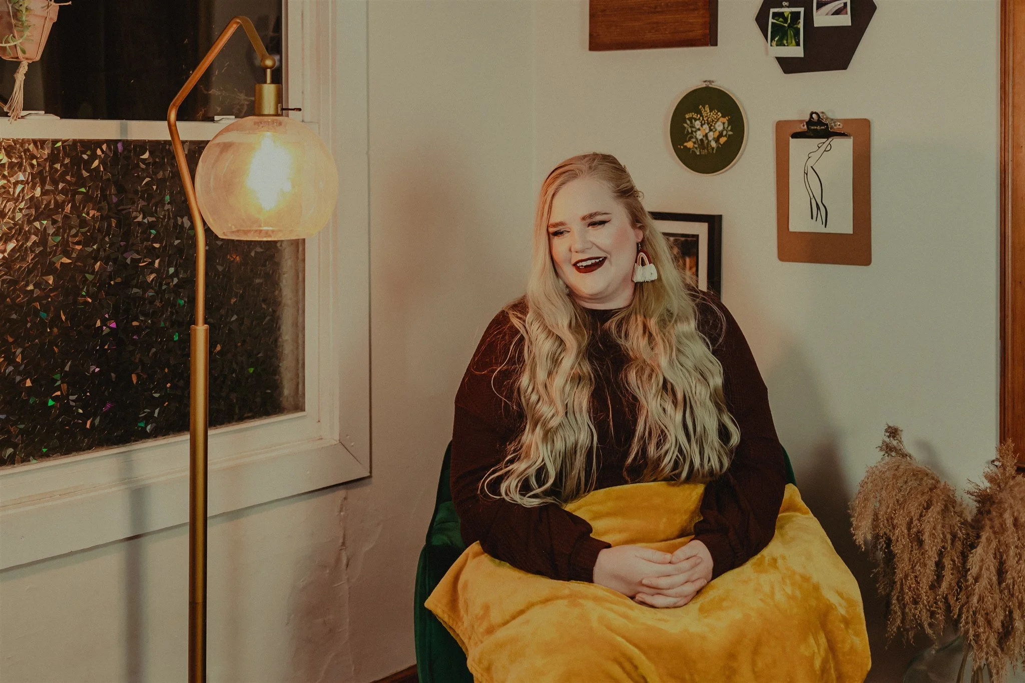 A woman with long wavy blonde hair and dark lipstick sitting on a green chair, smiling and holding a yellow blanket in her lap, in a cozy room with wall art and a floor lamp nearby during a trauma informed photography session in Columbia, Missouri.