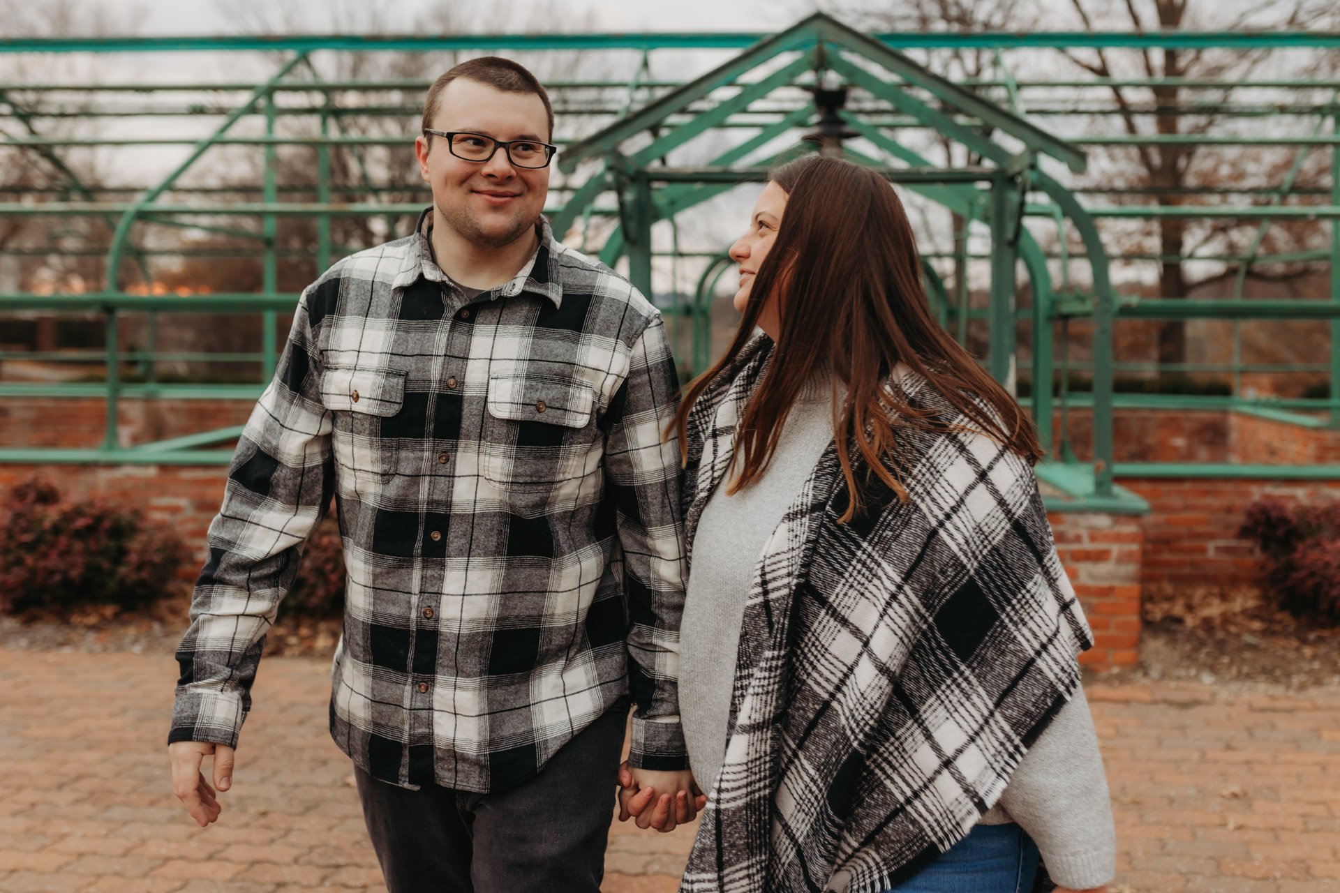 A couple holding hands and smiling at each other outdoors, with a greenhouse in the background at their engagement photography session at Queeny Park in STL.