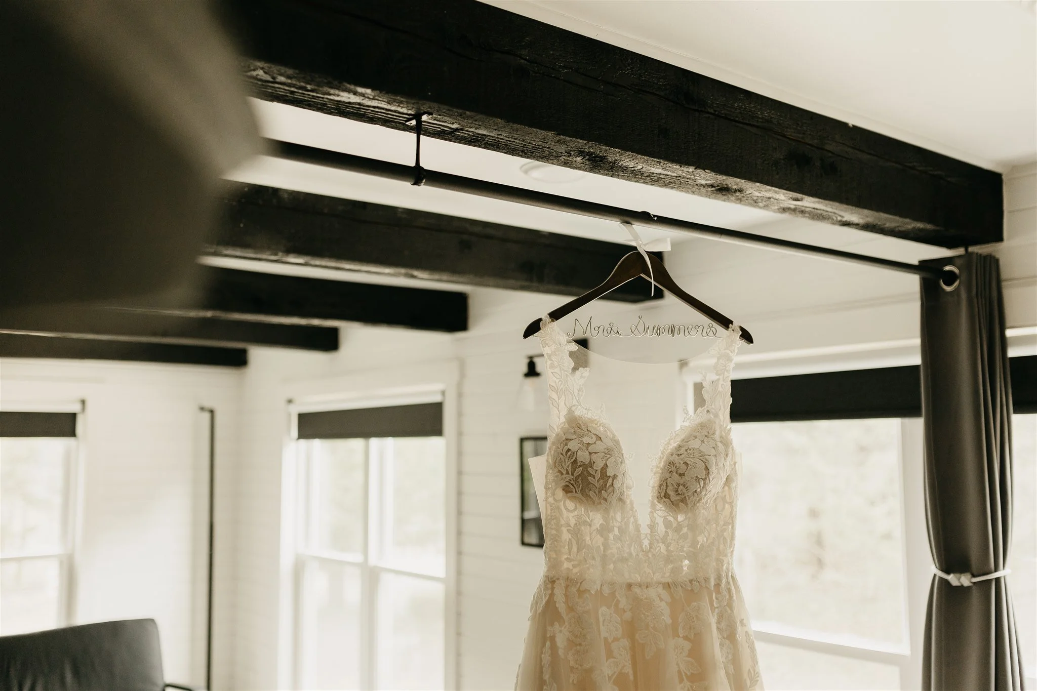 Wedding dress hanging on a wooden hanger inscribed with 'Mrs. Summer' in a bright room with windows and black curtain rods at Rolla, MO rustic wedding venue, The Village.