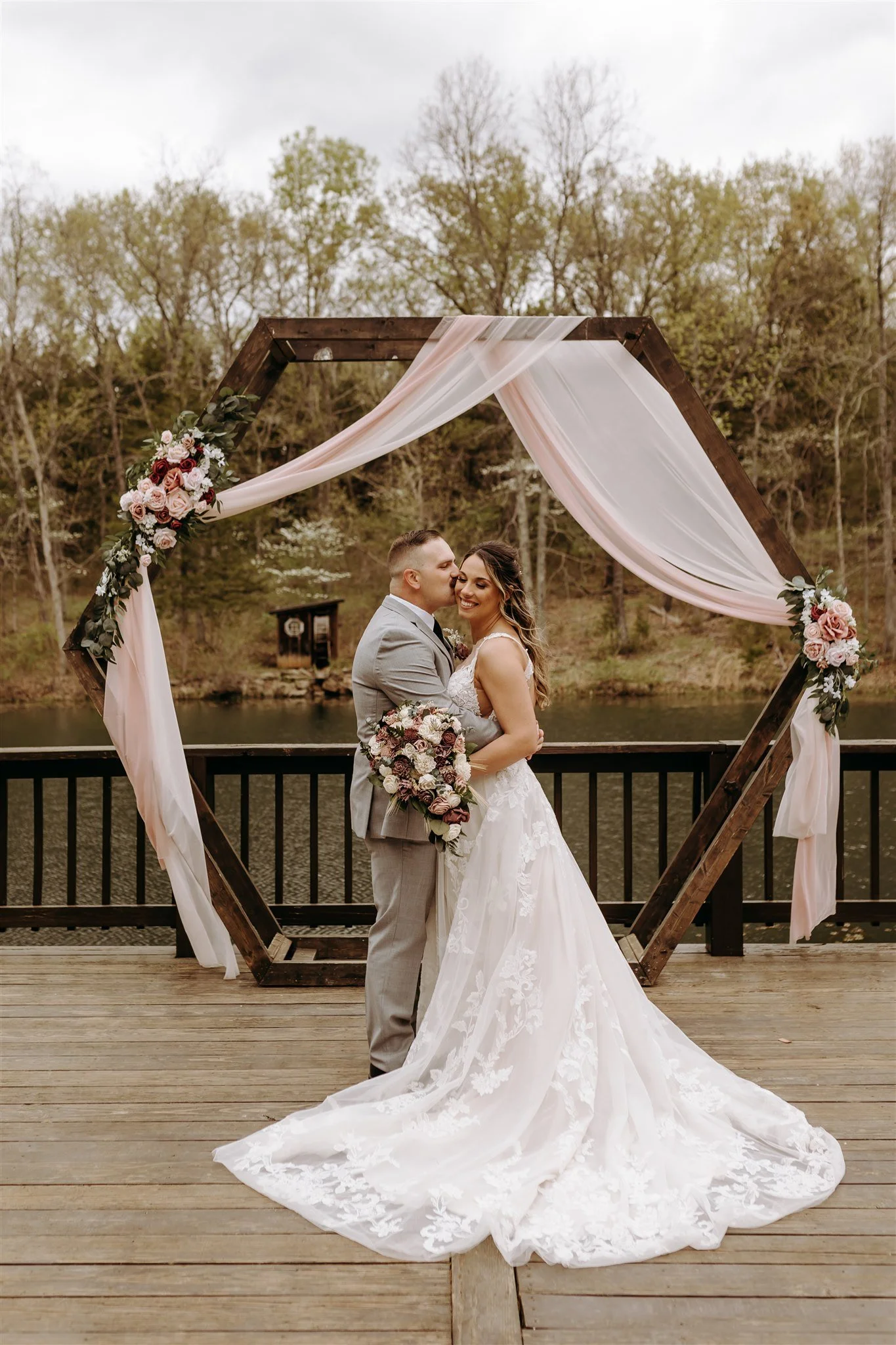 A newlywed couple in wedding attire standing on a wooden deck near a lake at Rolla, MO rustic wedding venue, The Village. Photos by Columbia, MO Photographer Liv Strange Photography.