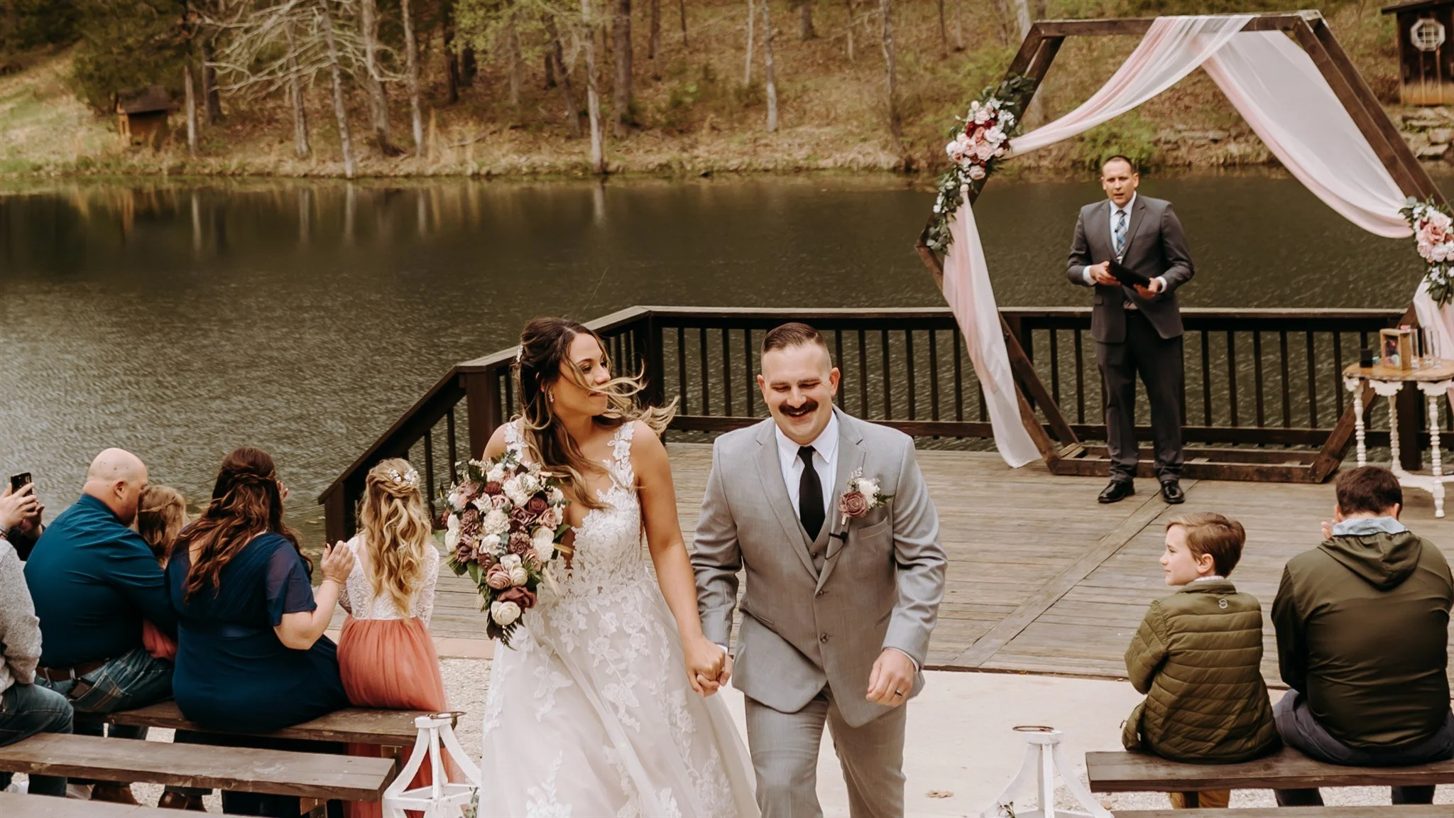 A wedding ceremony by a lake with the bride and groom holding hands and smiling, surrounded by seated guests at Rolla, Missouri rustic wedding venue, The Village. Photos by Columbia, MO Photographer Liv Strange Photography.