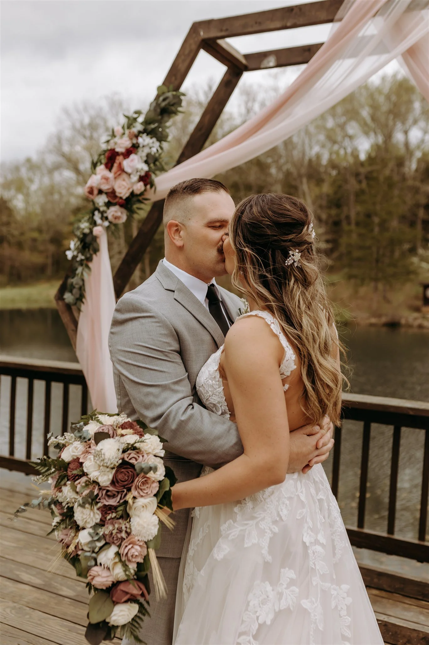 A newlywed couple sharing a kiss during their outdoor wedding ceremony on a wooden deck. The groom is dressed in a gray suit and the bride is in a white lace wedding gown holding a large floral bouquet at Rolla, MO rustic wedding venue, The Village.