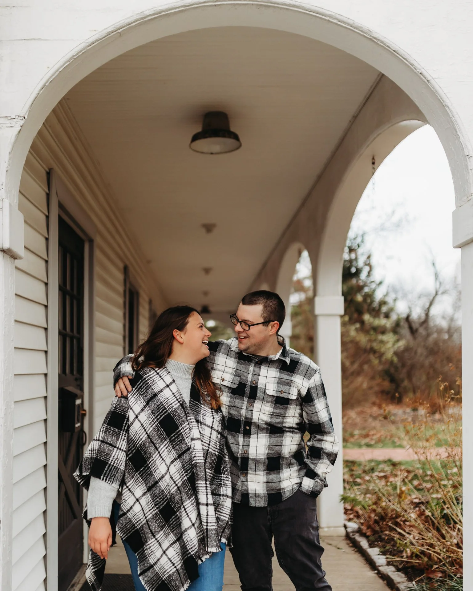 A young couple stands on a porch, smiling and looking at each other, dressed in matching plaid shirts and jackets during fall at their engagement photography session at Queeny Park in St. Louis, MO