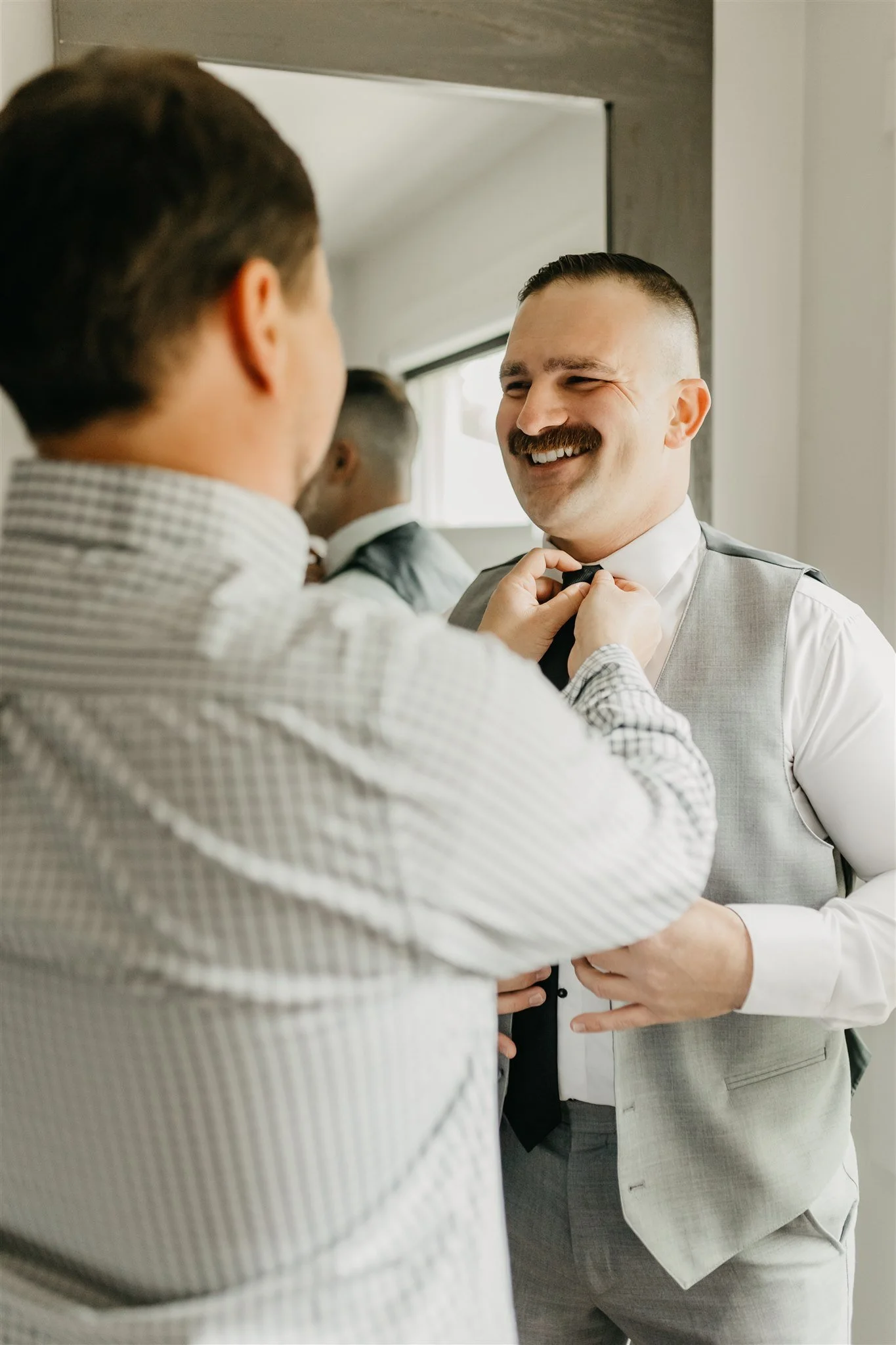 A man in a gray vest and white shirt smiling as another man helps him adjust his black tie in front of a mirror at Rolla, Missouri rustic wedding venue, The Village. Photos by Columbia, MO Photographer Liv Strange Photography.
