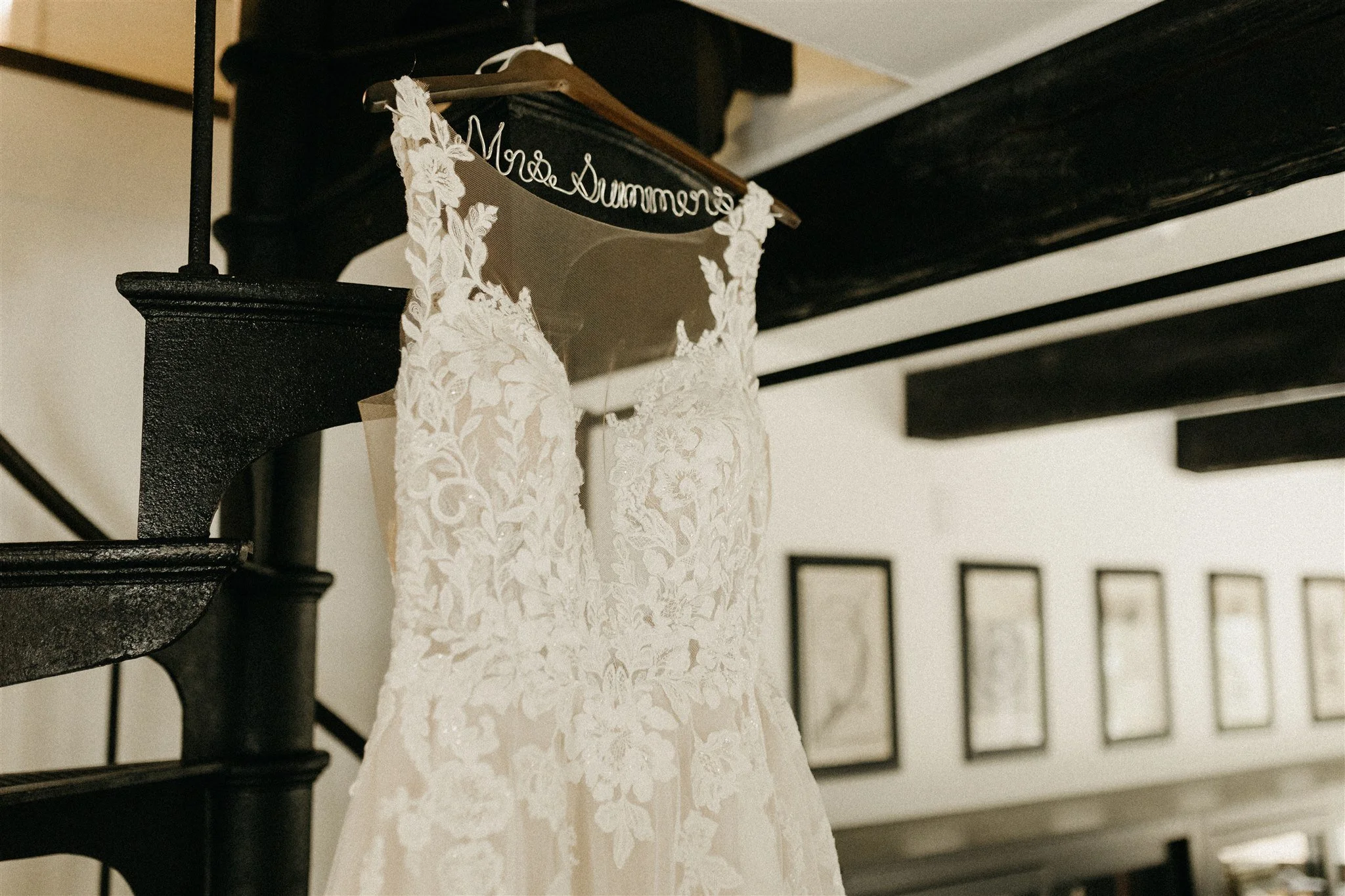 Wedding dress with lace details hanging on a wooden hanger with the words 'Mrs. Summer' embroidered in cursive, displayed on a spiral staircase in a room with framed pictures on the wall at Rolla, MO rustic wedding venue, The Village.