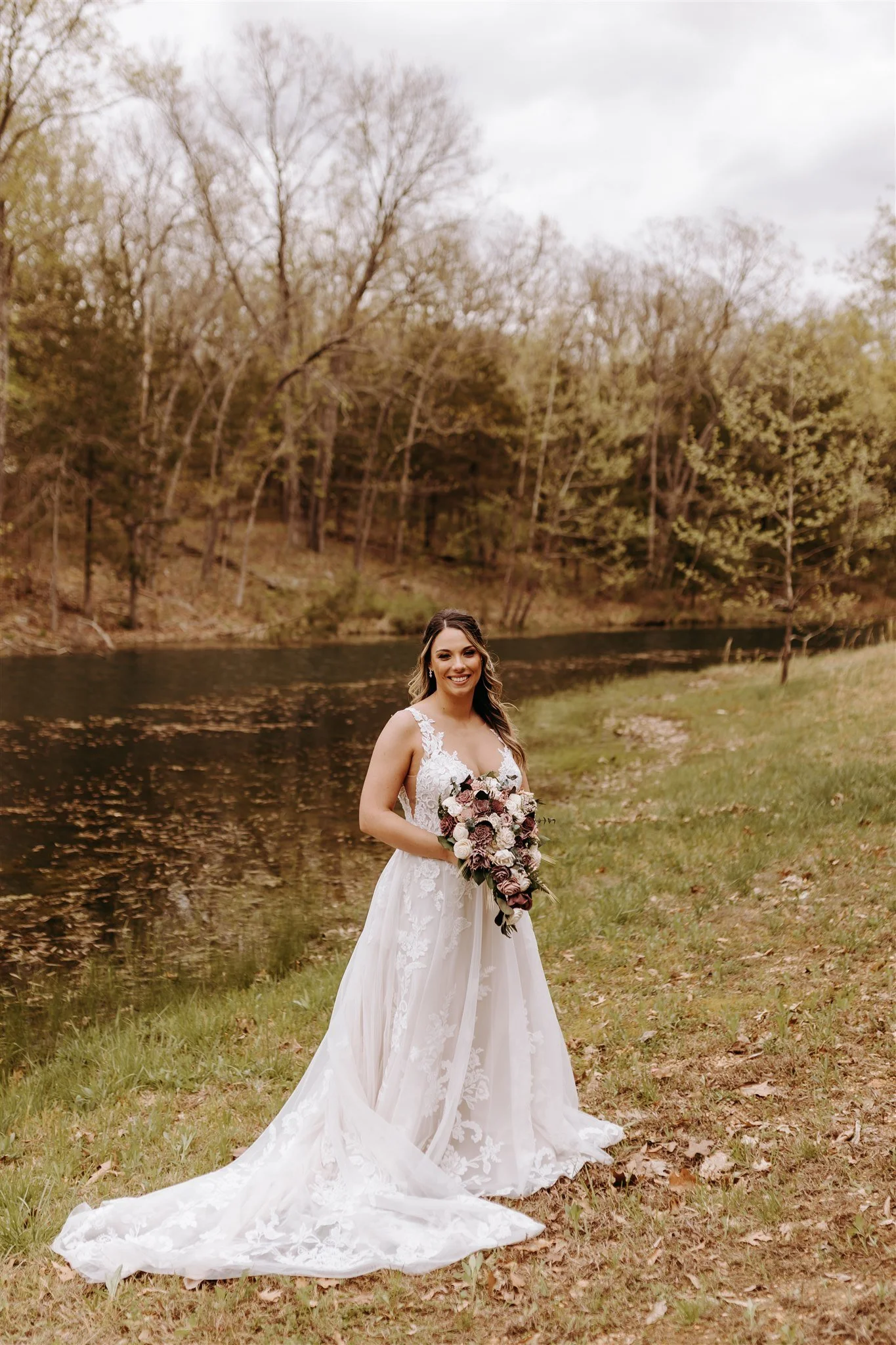 A bride standing on a grass bank next to a river, holding a large bouquet of flowers, wearing a white lace wedding dress, with trees and overcast sky in the background at Rolla, MO rustic wedding venue, The Village.