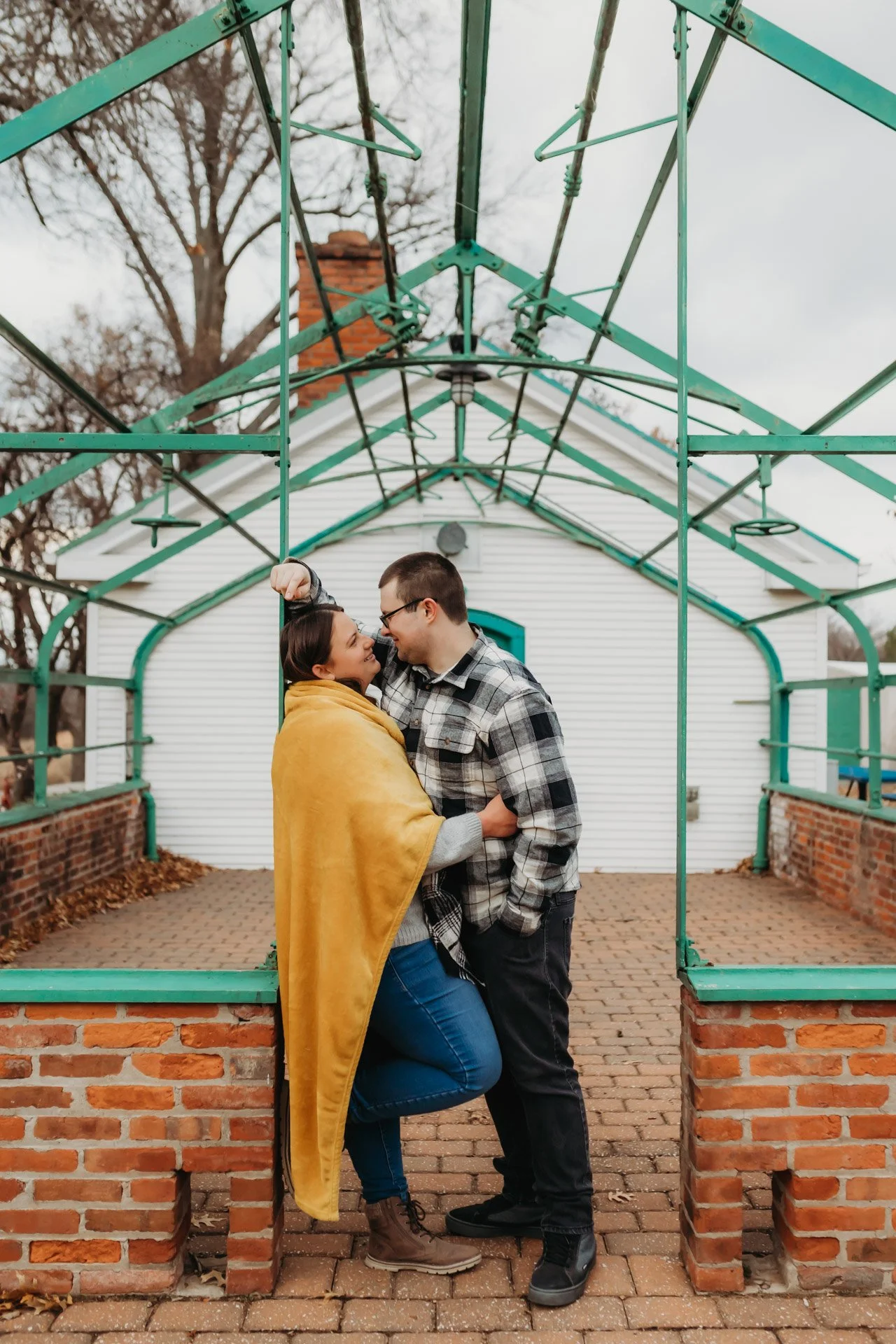 A couple standing close together, gazing into each other's eyes, under a green metal framework in front of a white house, with the woman wrapped in a yellow blanket at their engagement photography session at Queeny Park in STL.