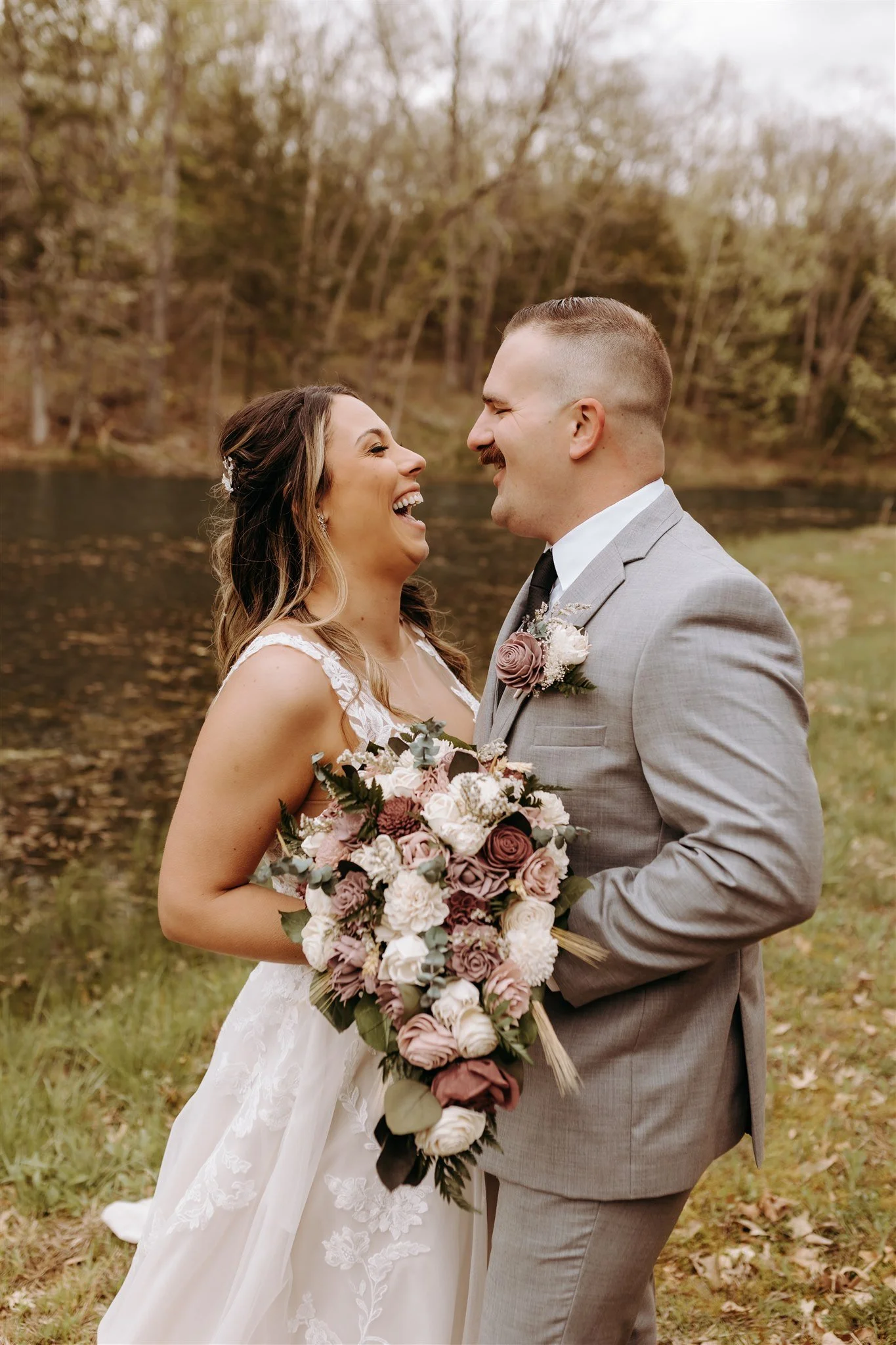 A bride and groom laughing while holding a bouquet of flowers outdoors near a lake, with trees in the background at Rolla, MO rustic wedding venue, The Village. Photos by Columbia, MO Photographer Liv Strange Photography.