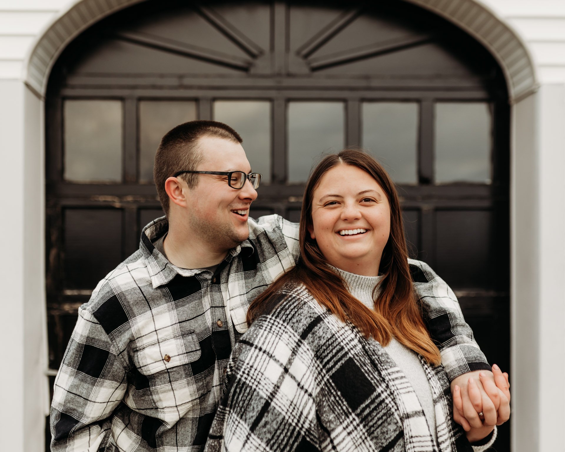A happy couple stands outdoors, smiling and looking at each other, with the woman in front and the man behind her, wearing casual plaid shirts, in front of a black garage door at their engagement photography session at Queeny Park in STL.