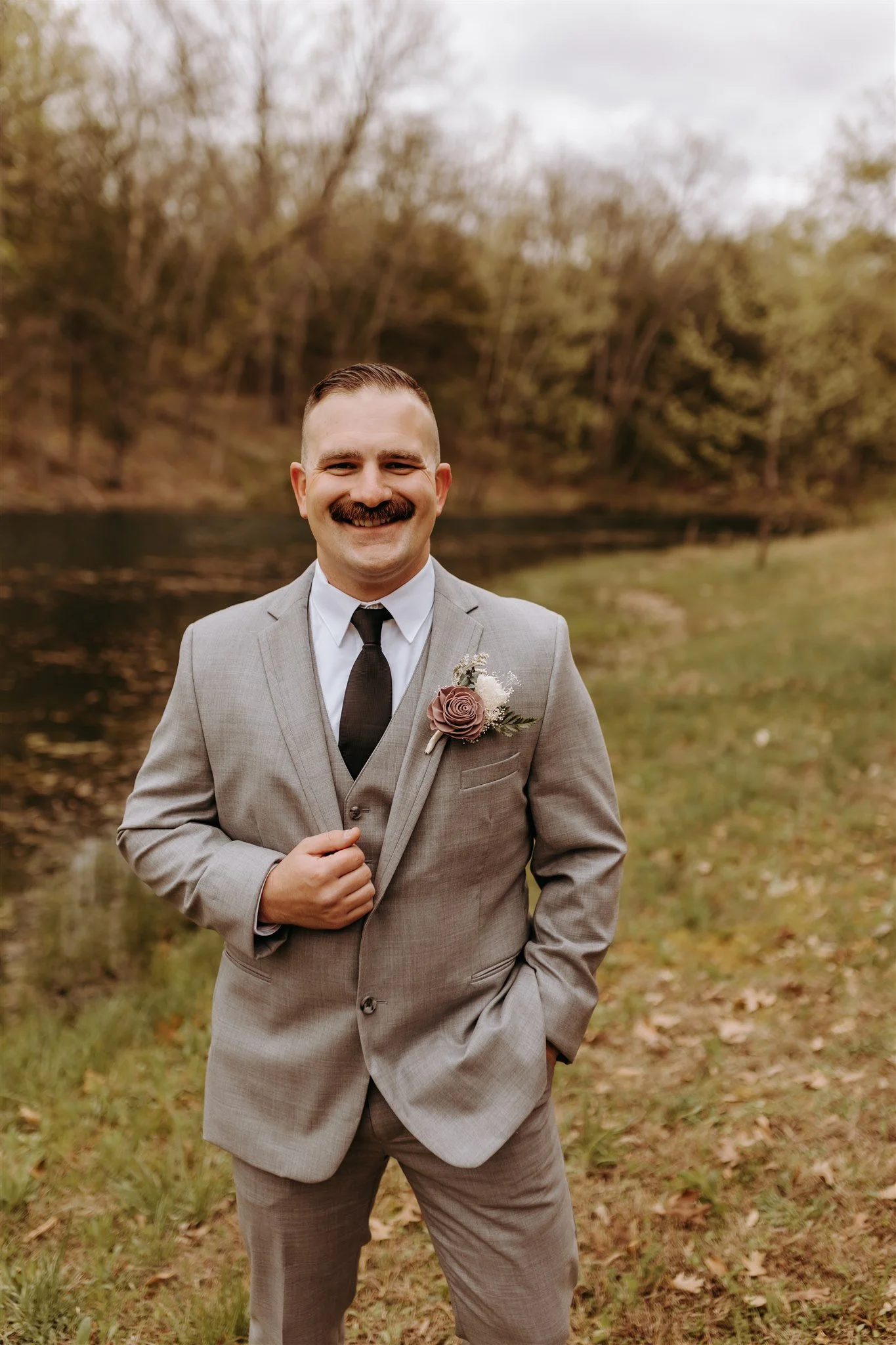 A groom with a mustache dressed in a light gray suit, white shirt, and black tie stands outdoors near a body of water and trees, smiling at the camera at Rolla, MO rustic wedding venue, The Village. Photos by Columbia, MO Photographer.