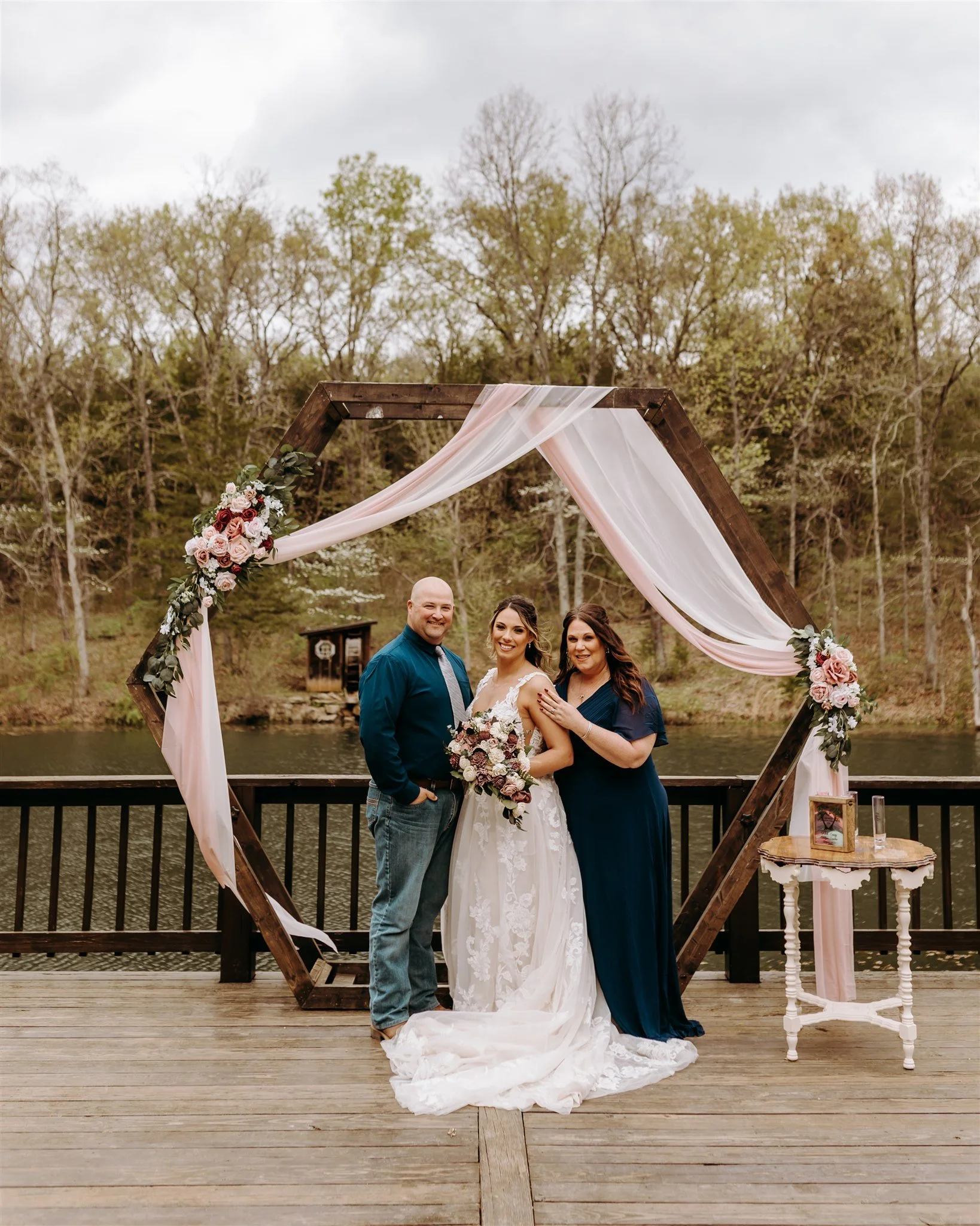 A wedding couple and a woman standing under an arch decorated with pink and white flowers and drapes on a wooden deck by a lake surrounded by trees at Rolla, Missouri rustic wedding venue, The Village. Photos by Columbia, MO Photographer Liv Jones.