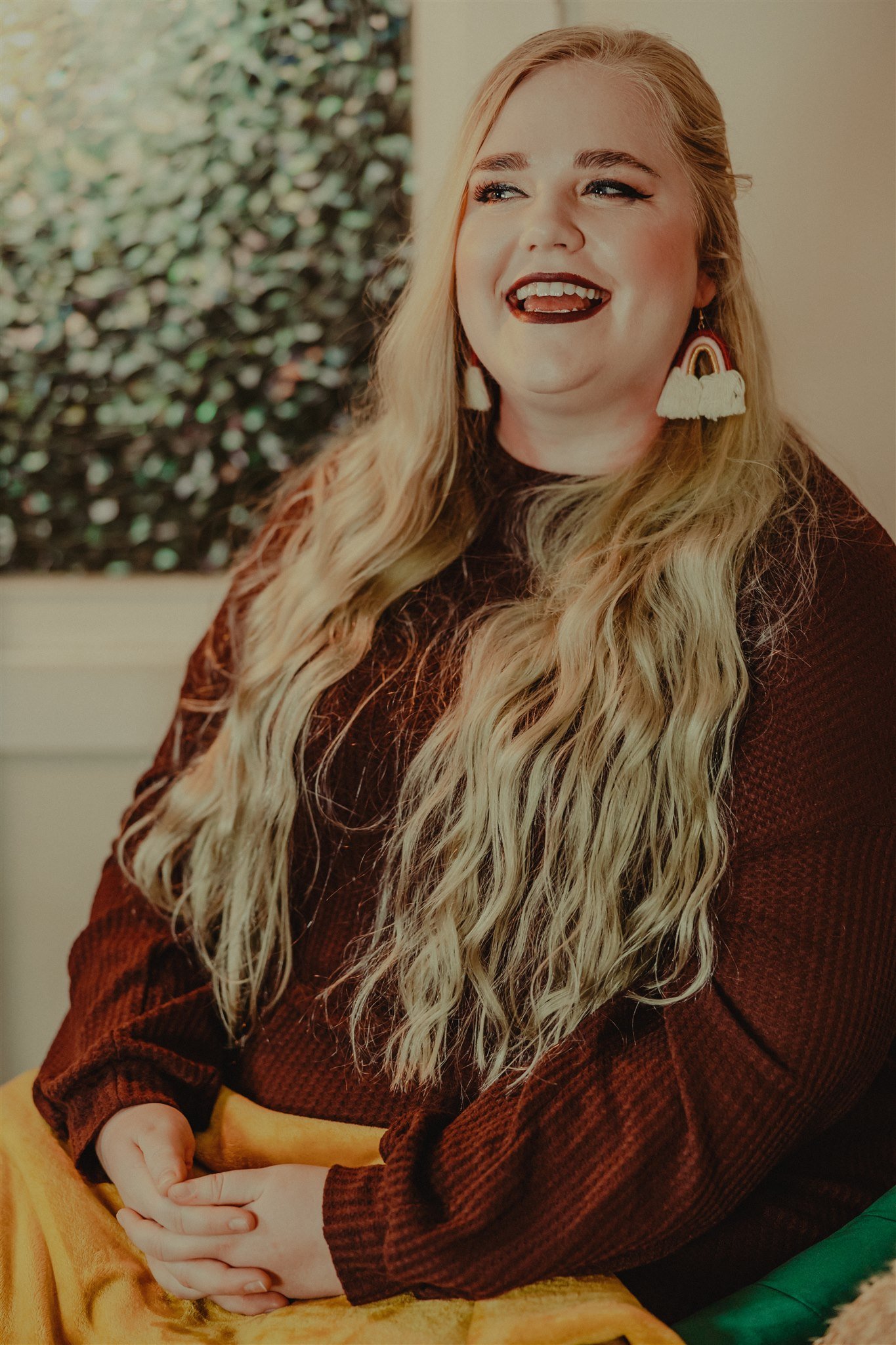 A woman with long, wavy blonde hair wearing a maroon sweater and rainbow earrings, smiling and looking to the side indoors during a trauma informed photography session in Columbia, Missouri.