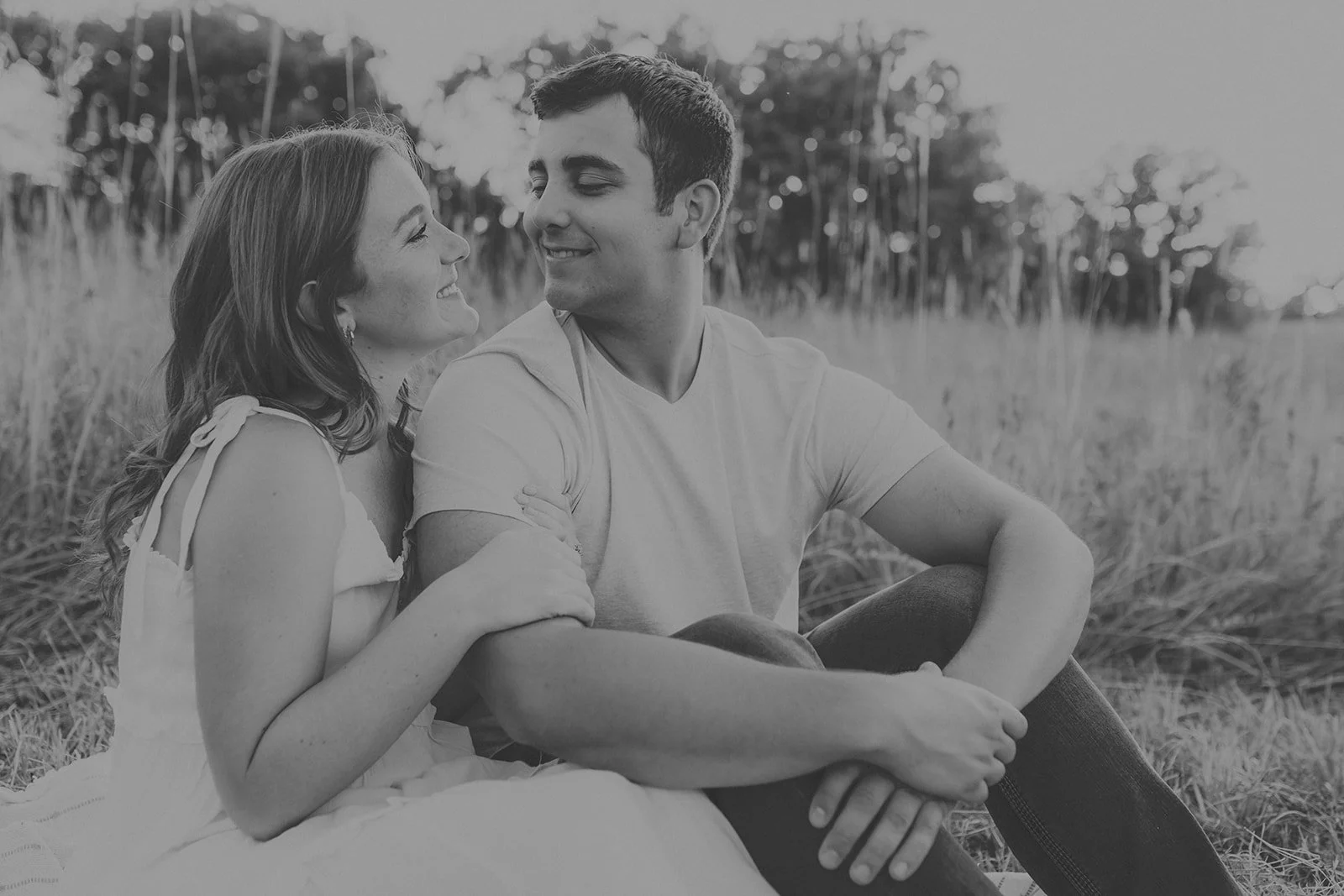 An engaged couple sitting in a grassy field, smiling and looking at each other, with trees in the background during their couples photography session at Rock Bridge State Park in Columbia, MO.