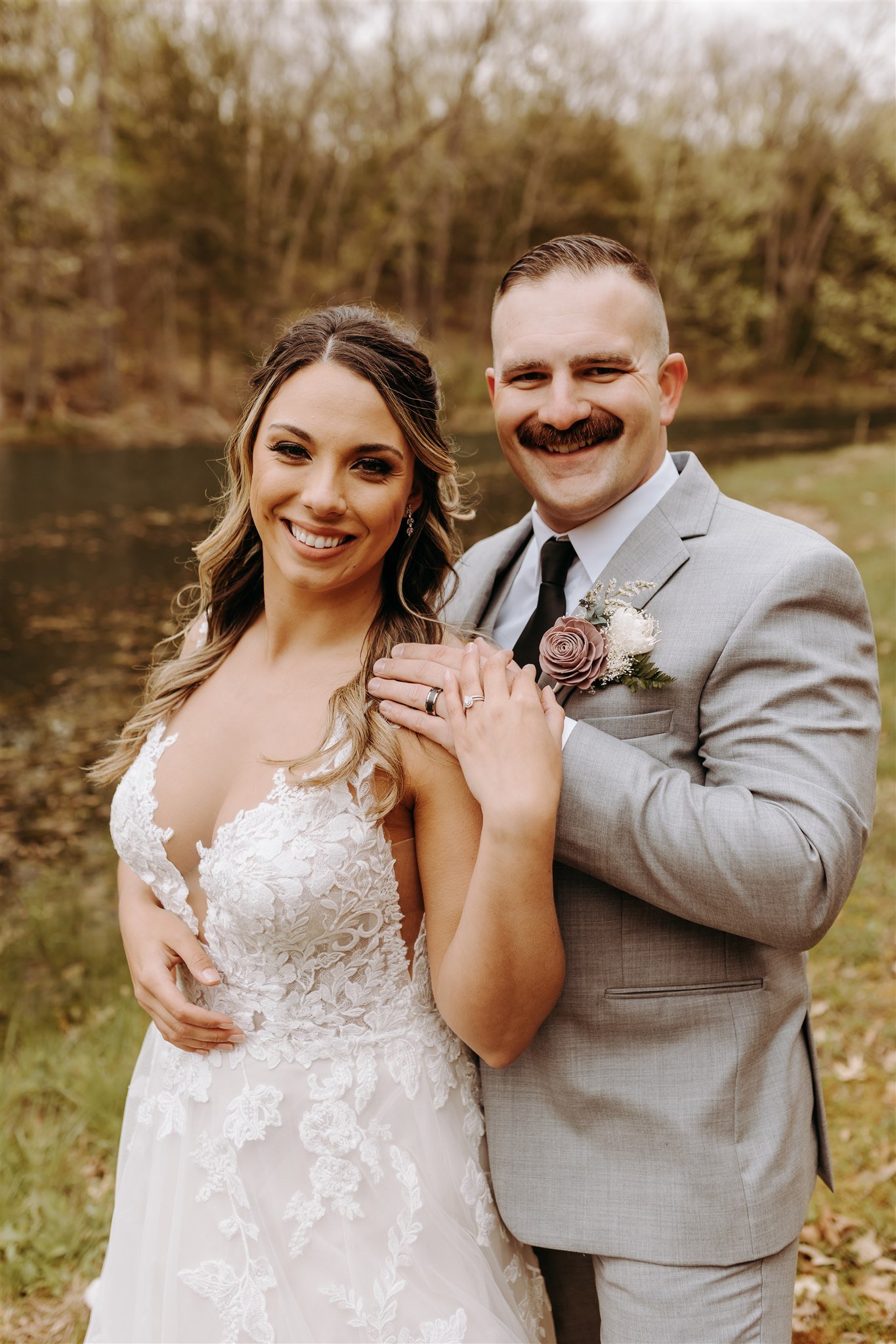 A smiling bride and groom in wedding attire outdoors with trees and a body of water in the background at Rolla, MO rustic wedding venue, The Village. Photos by Columbia, MO Photographer Liv Strange Photography.