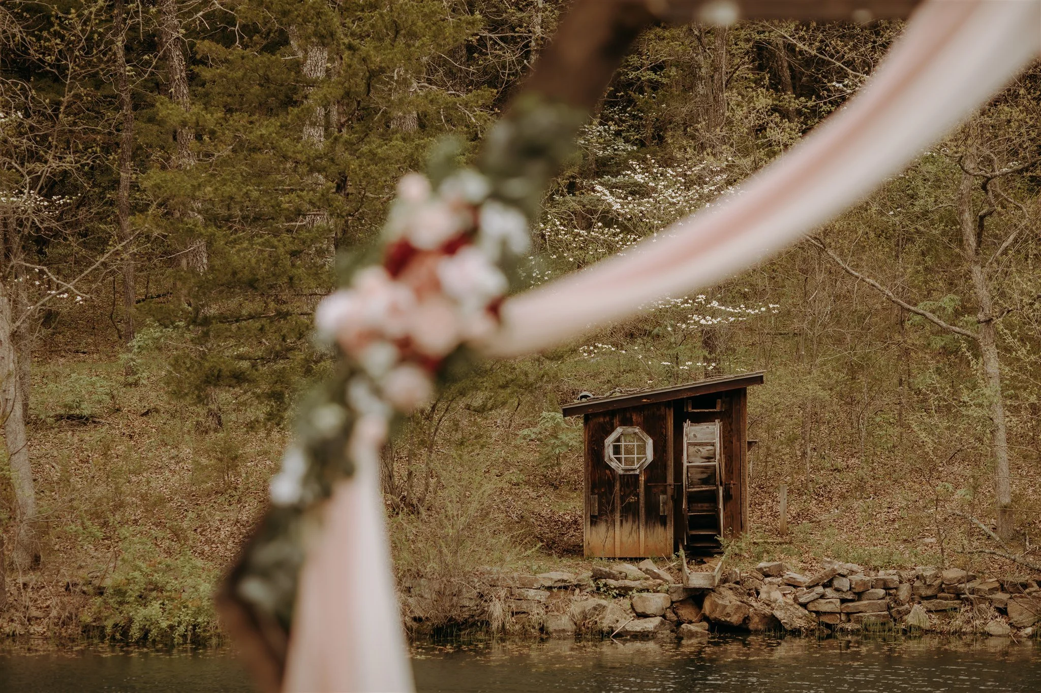 Blurred floral decoration in the foreground, with a small rustic wooden shed on the water's edge and trees in the background at Rolla, MO rustic wedding venue, The Village.