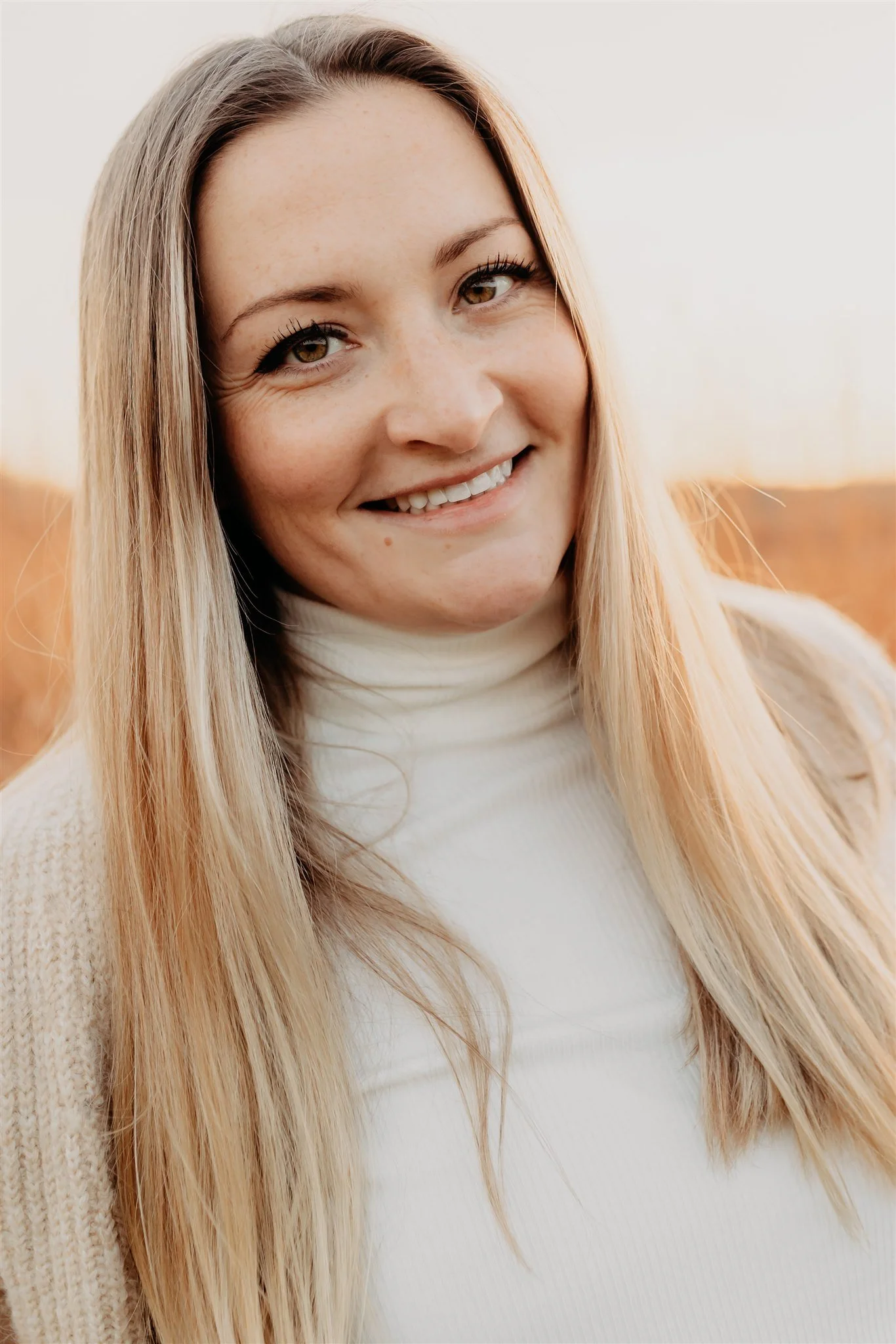 Close-up of a young woman with long blonde hair, smiling, wearing a cream turtleneck sweater, outdoors during her Columbia, MO branding photography session for her small business.