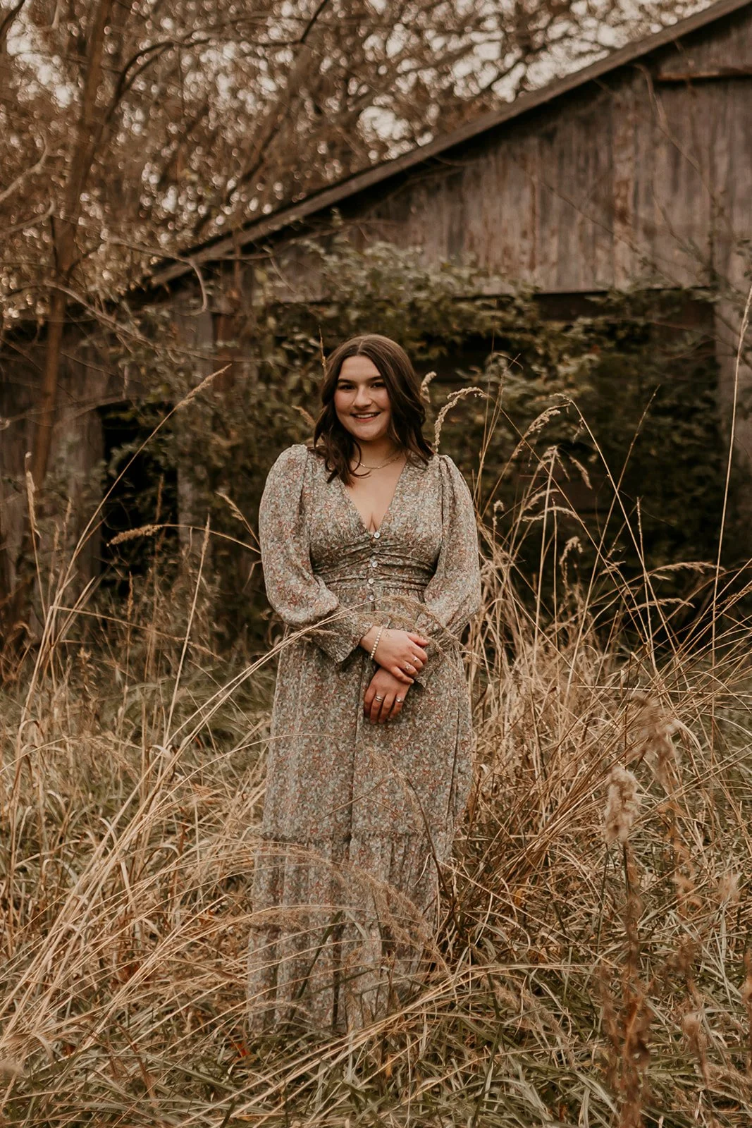 A young woman with shoulder-length dark hair in a floral dress standing in a field of tall, dry grass with an old wooden barn and trees in the background.