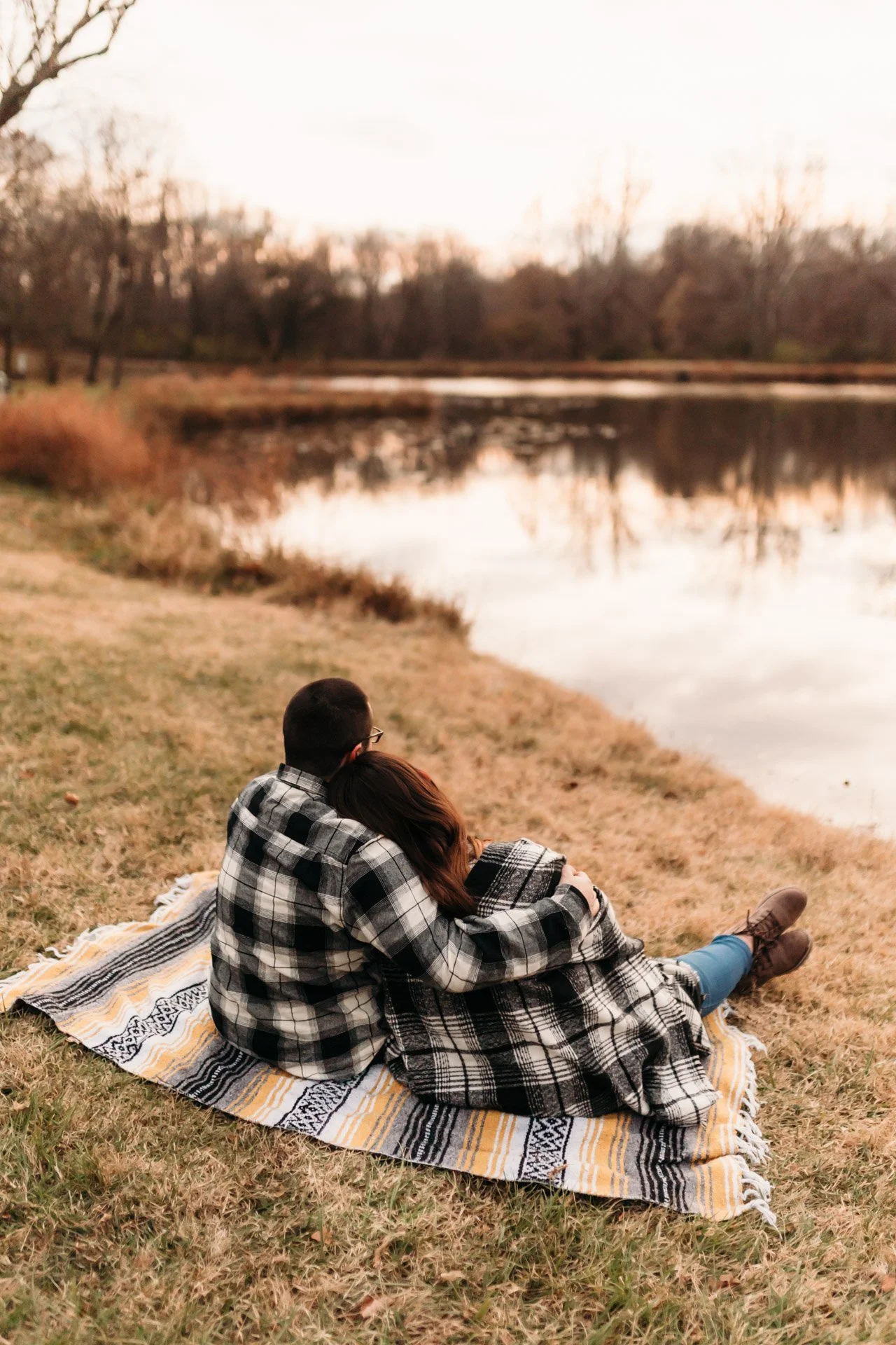 A couple sitting on a blanket by a lake, embracing and enjoying the sunset in a fall setting at their engagement photography session at Queeny Park in STL.