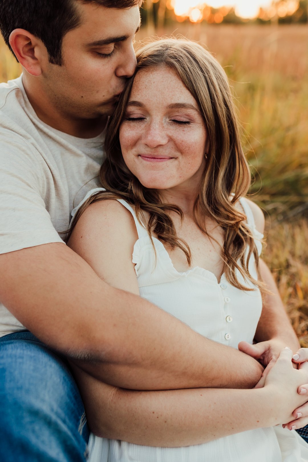 A young man kissing a young woman on the forehead while sitting outside at sunset. The woman is smiling with her eyes closed, and they are embracing each other during their engagement photography session at Rock Bridge State Park in Columbia, MO.