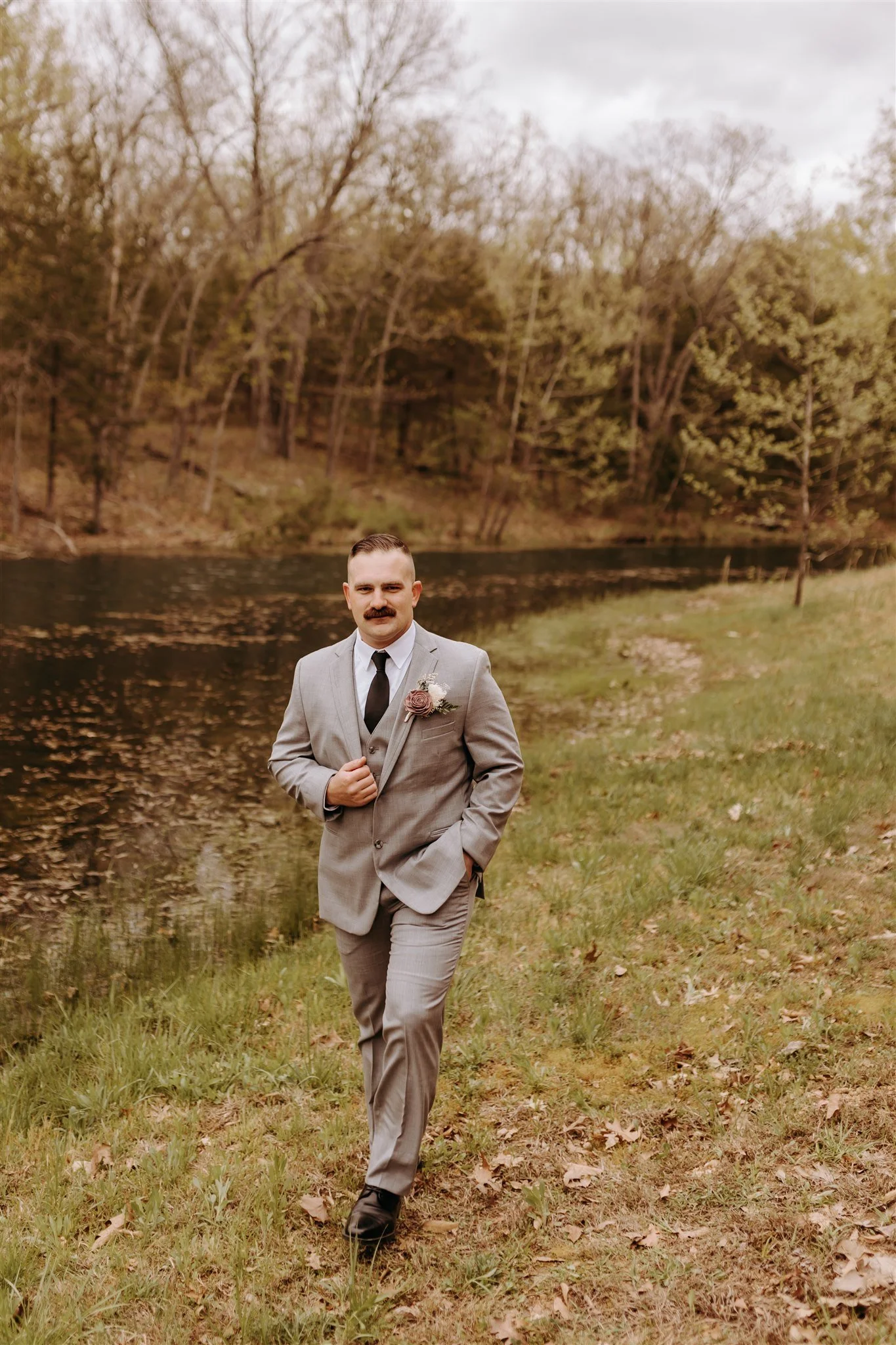 A groom in a gray suit standing outdoors near a lake, with trees in the background, during overcast weather at Rolla, MO rustic wedding venue, The Village. Photos by Columbia, MO Photographer Liv Strange Photography.