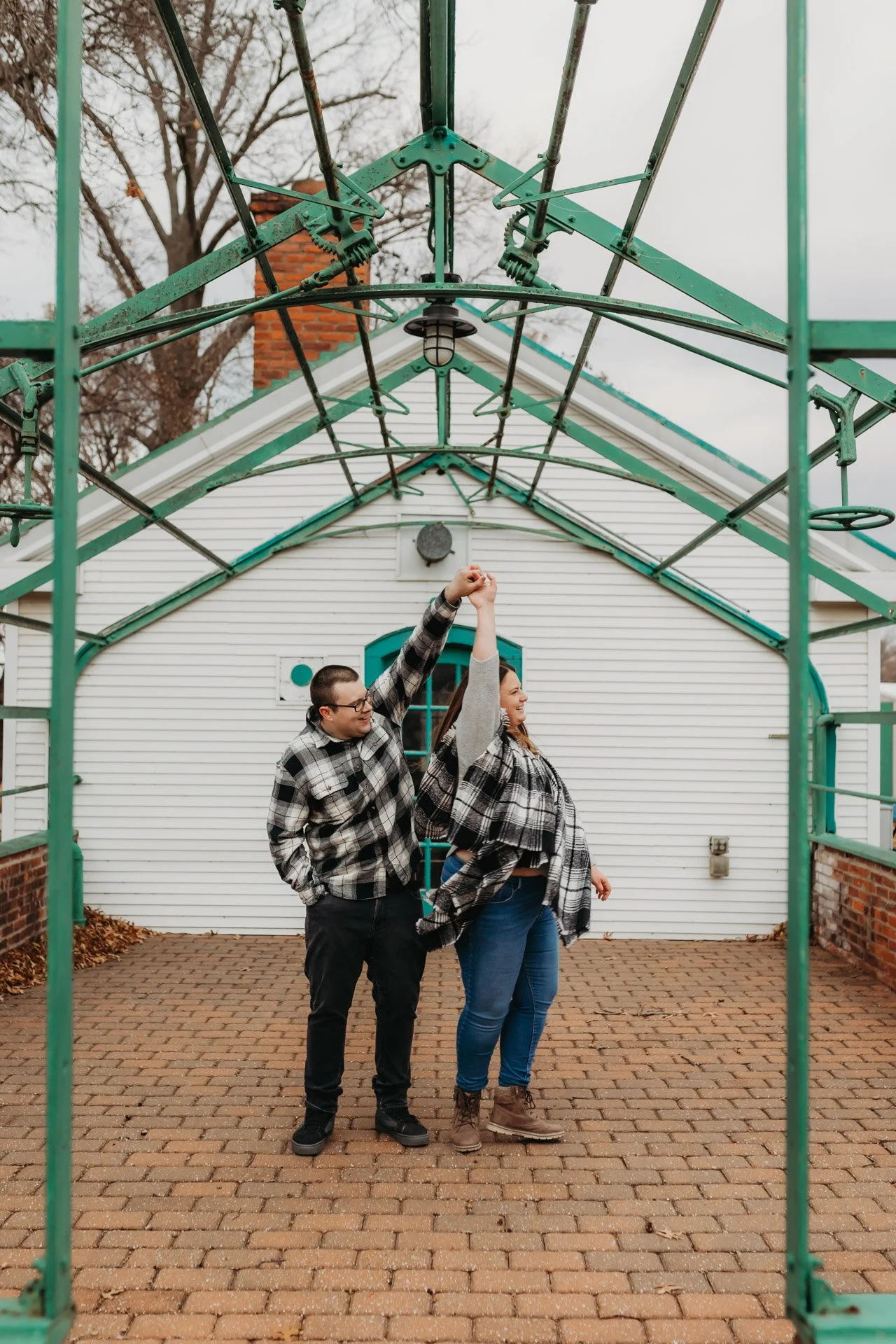 A man and a woman standing on brick ground, holding hands raised, under a green metal arbor frame, with a white house with blue trim in the background at their engagement photography session at Queeny Park in STL.