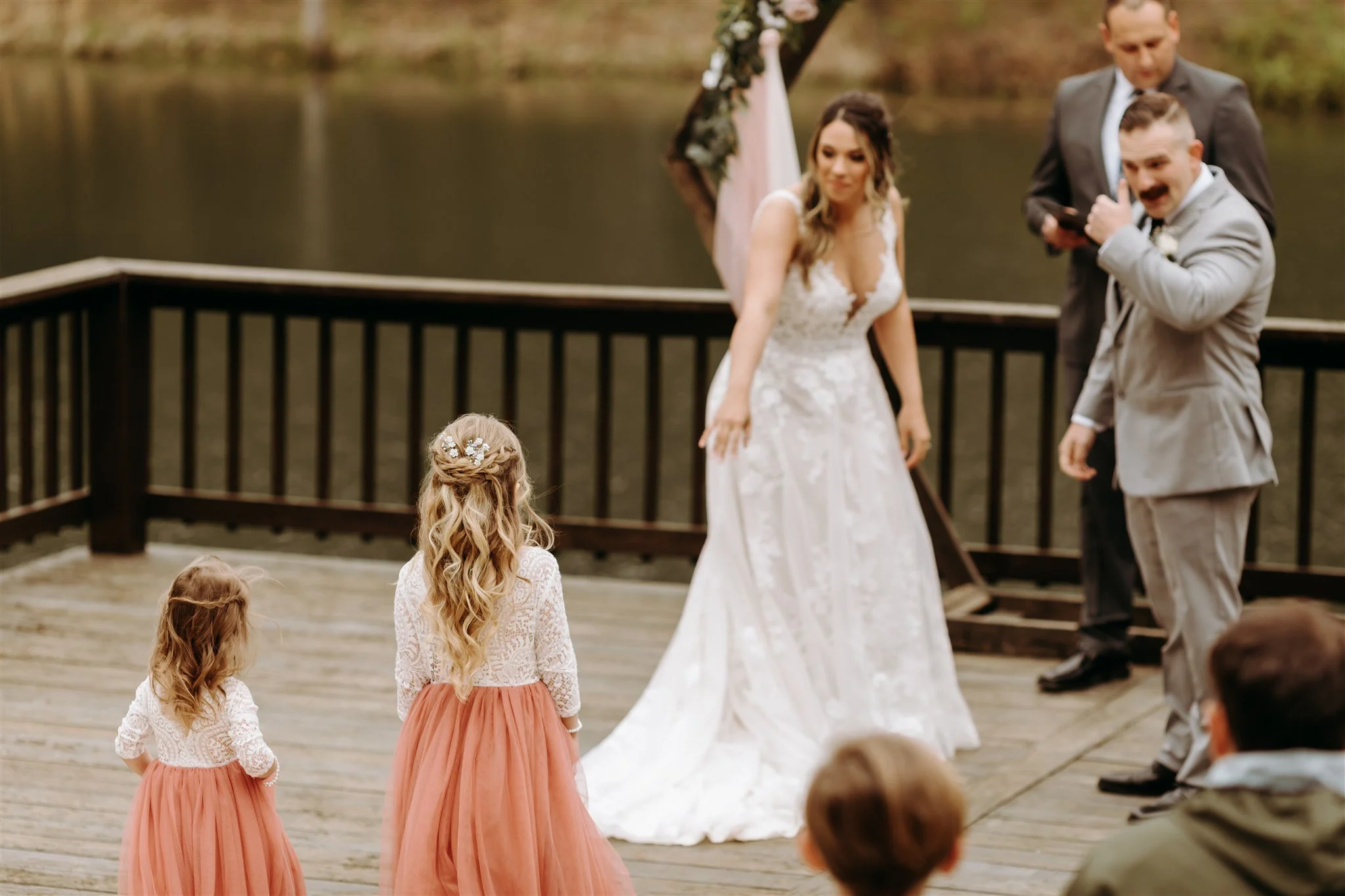 Bride, two young flower girls in matching lace tops and peach skirts, groom in a light gray suit, officiant in dark suit, during wedding ceremony. Guests watching by a lake at Rolla, Missouri rustic wedding venue, The Village.