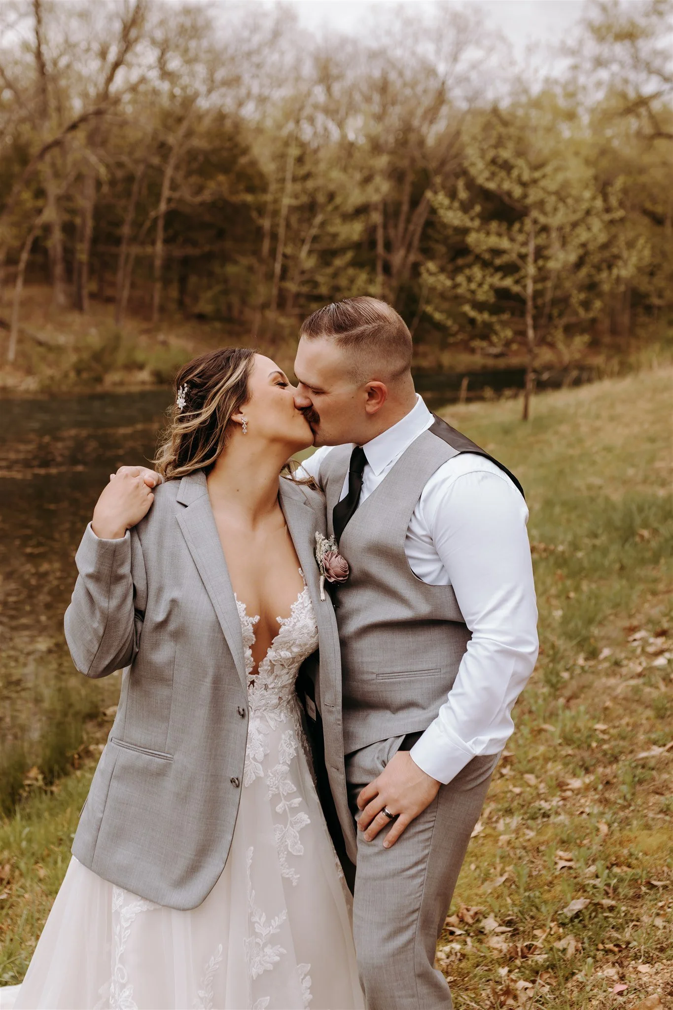 A newlywed couple sharing a kiss outdoors in a natural setting with trees and water in the background. The bride is wearing a lace wedding gown and a gray blazer at Rolla, MO rustic wedding venue, The Village.