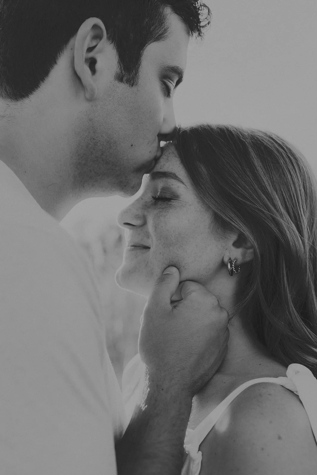A black and white photo of a couple sharing an intimate moment; the man is gently holding the woman's chin, with the woman smiling softly and eyes closed during their engagement photography session at Rock Bridge State Park in Columbia, MO.