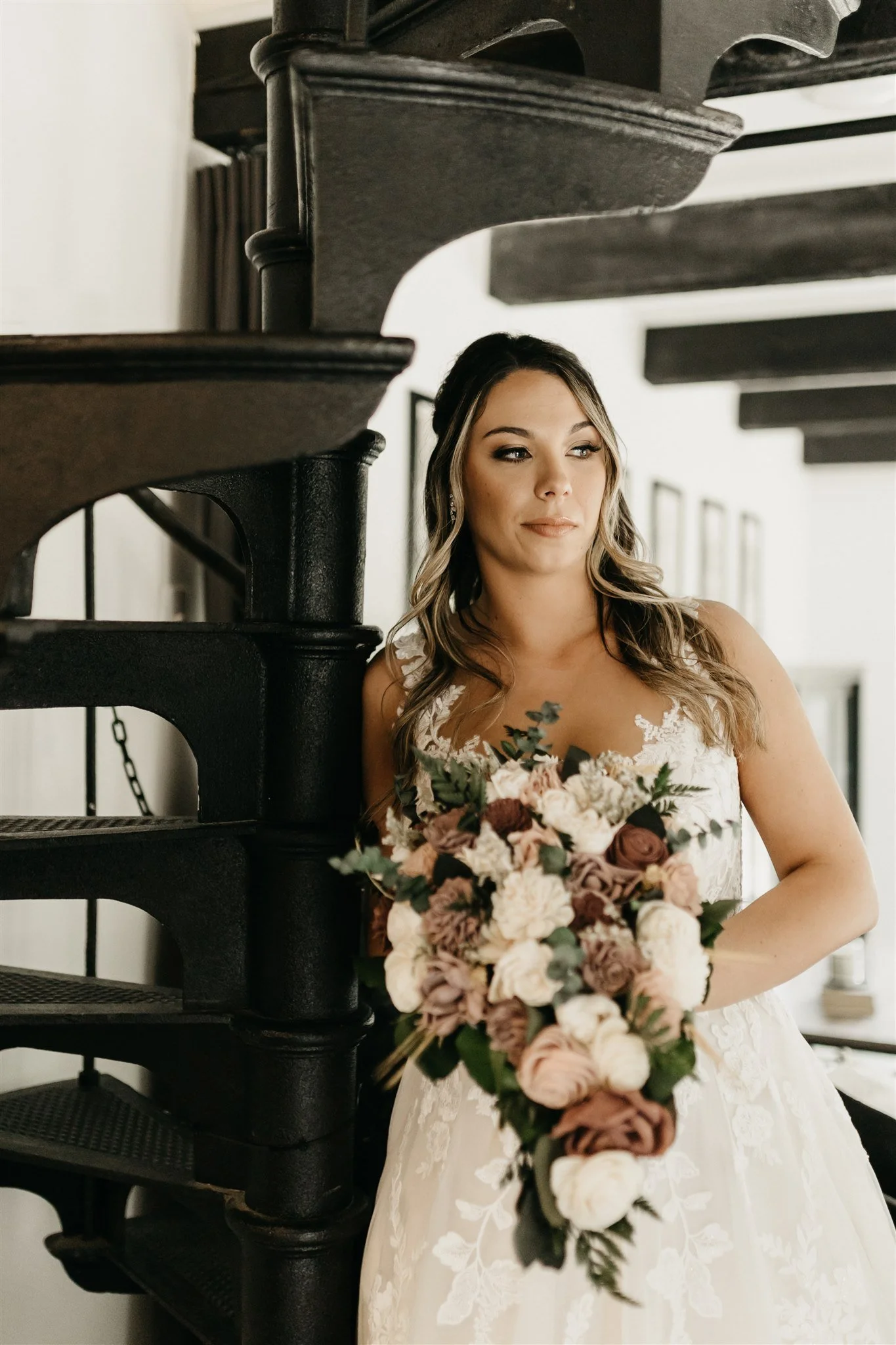 A bride in a white lace wedding dress holding a large bouquet, standing next to a black staircase in an indoor setting at Rolla, Missouri rustic wedding venue, The Village. Photos by Columbia, MO Photographer Liv Strange Photography.