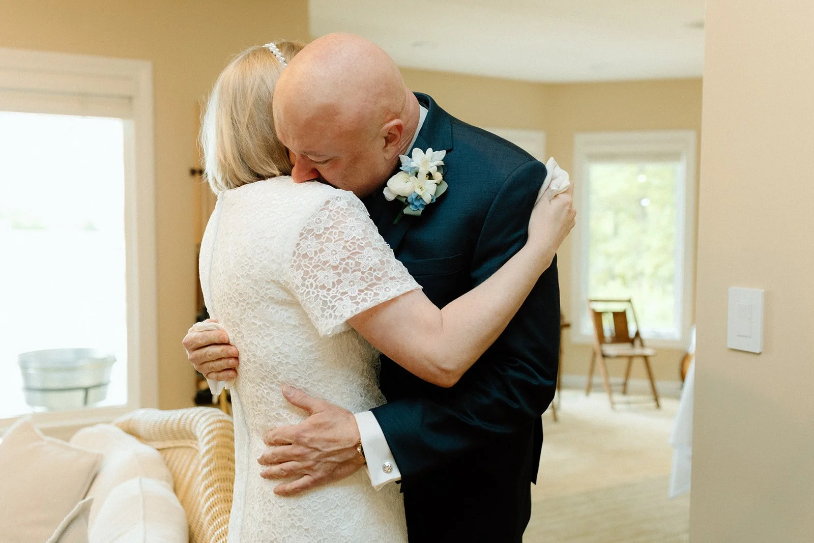 A man in a dark suit and a woman in a white lace dress sharing an emotional embrace, likely during a wedding ceremony, inside a bright room with large windows at an intimate backyard wedding in Columbia, MO.