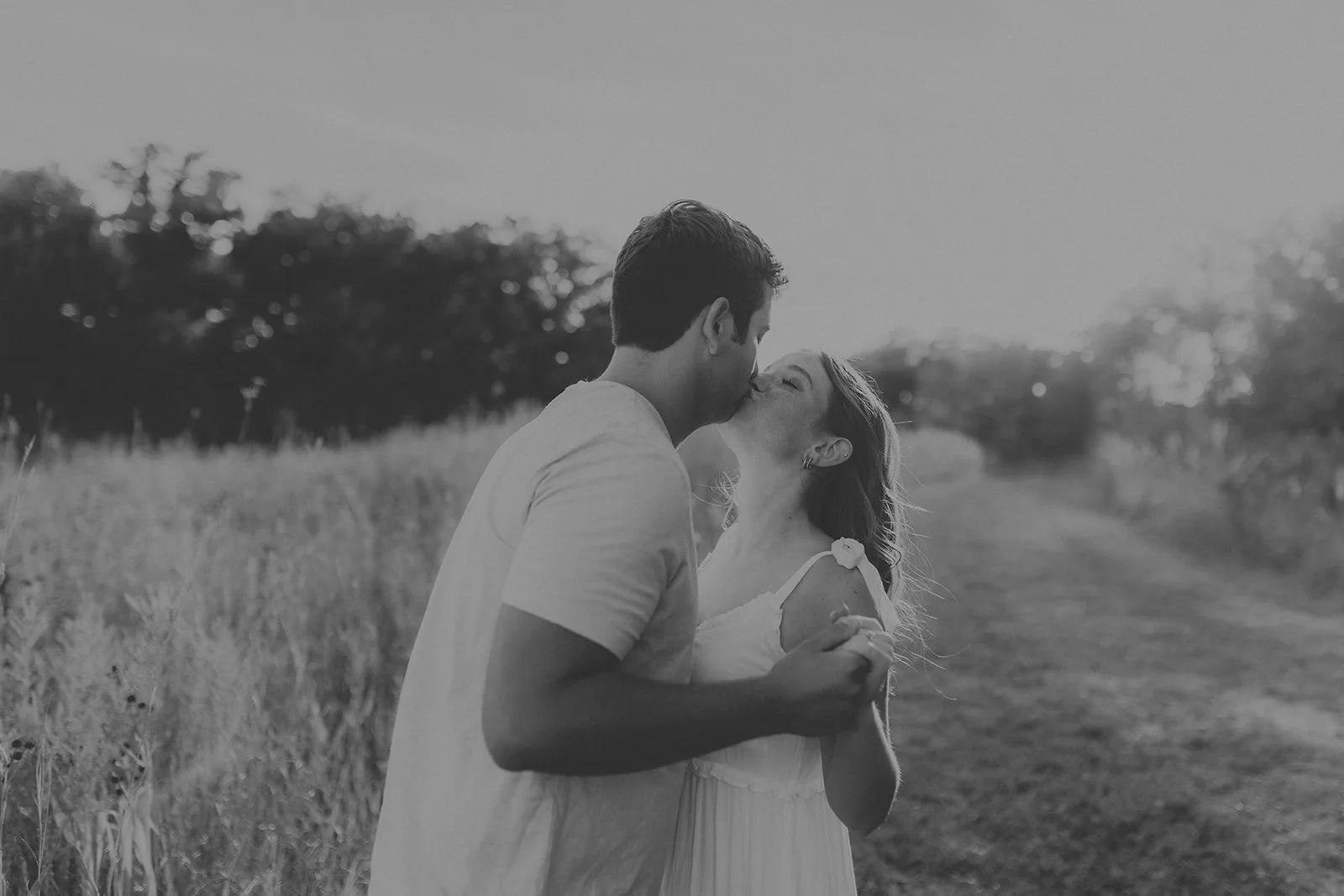 A black and white photo of a couple kissing in a field, with trees in the background and a dirt path during their engagement photography session at Rock Bridge State Park in Columbia, MO.