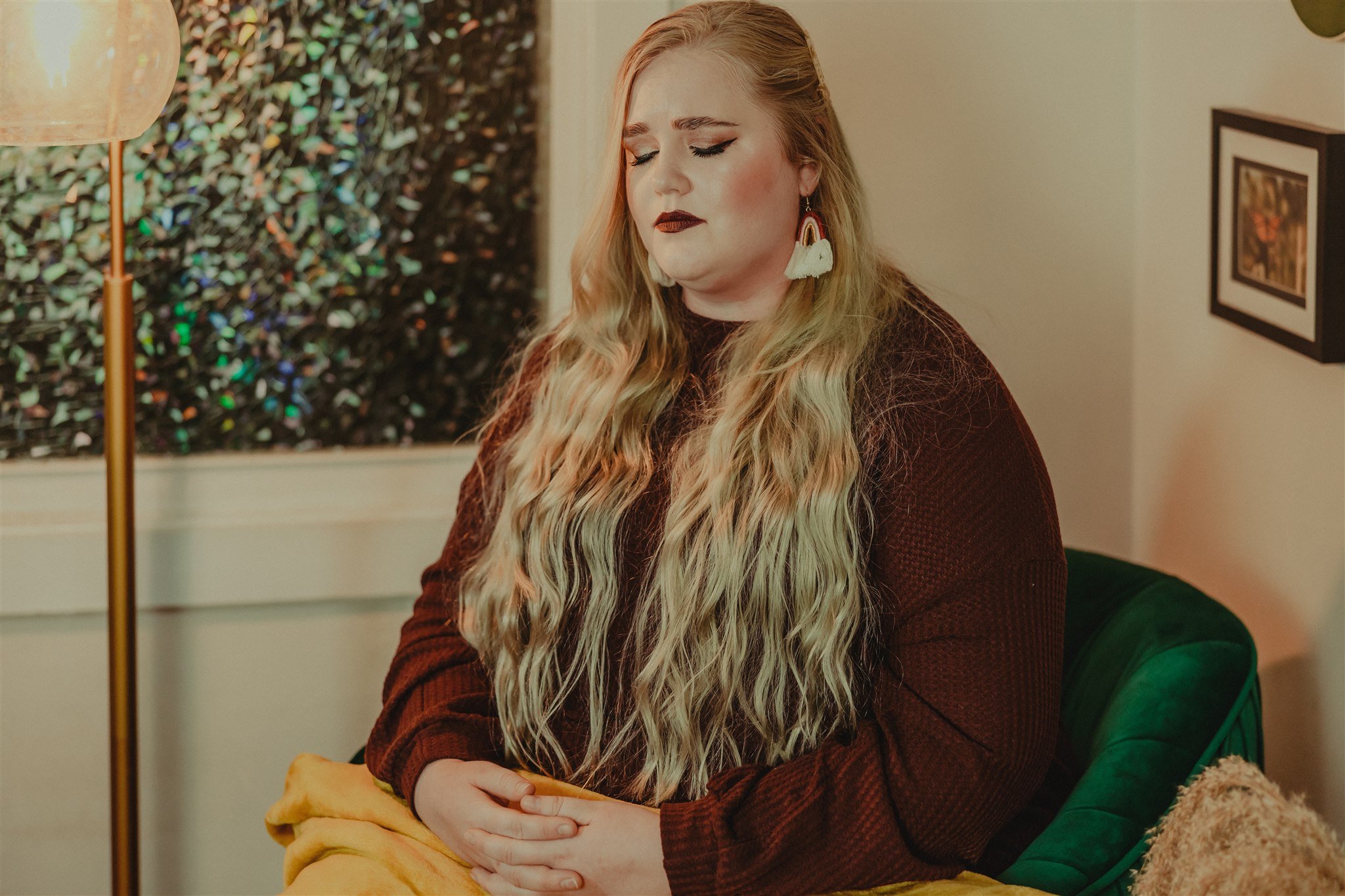 A woman with long wavy blonde hair and makeup, sitting in a room with a green velvet chair, a framed picture on the wall, and a lamp nearby during a trauma informed photography session in Columbia, Missouri.