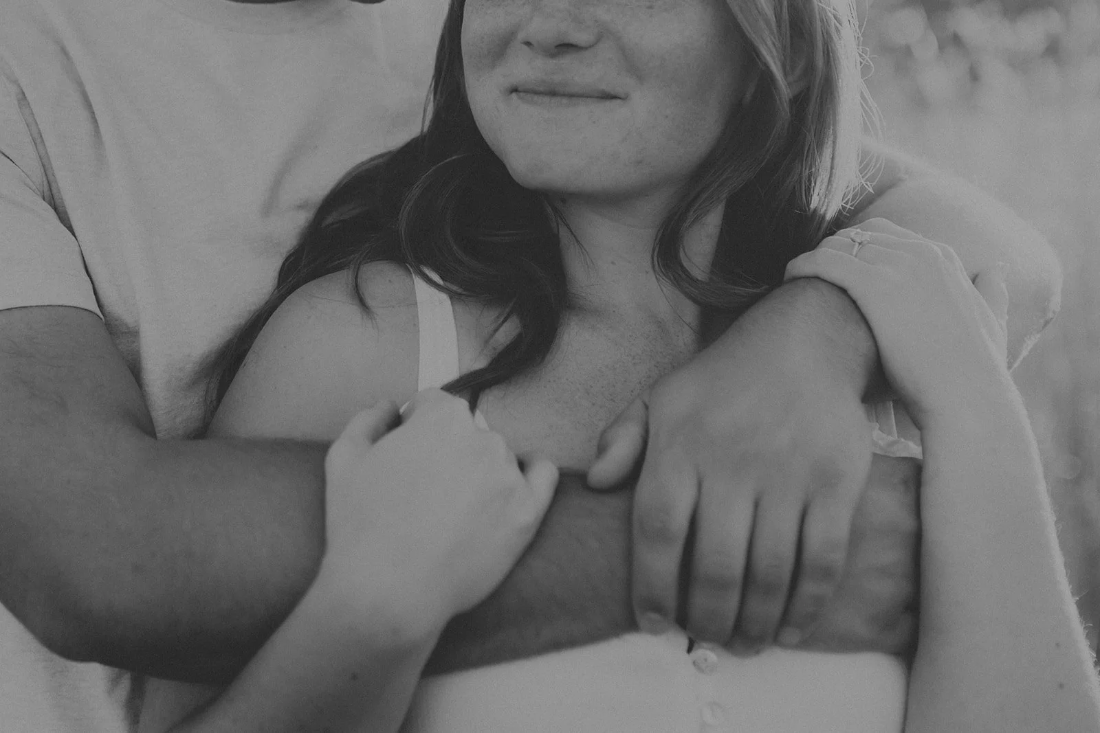 A woman with long dark hair is being embraced from behind, with her arms crossed over her chest and gentle hold of her hands during their engagement photography session at Rock Bridge State Park in Columbia, MO.