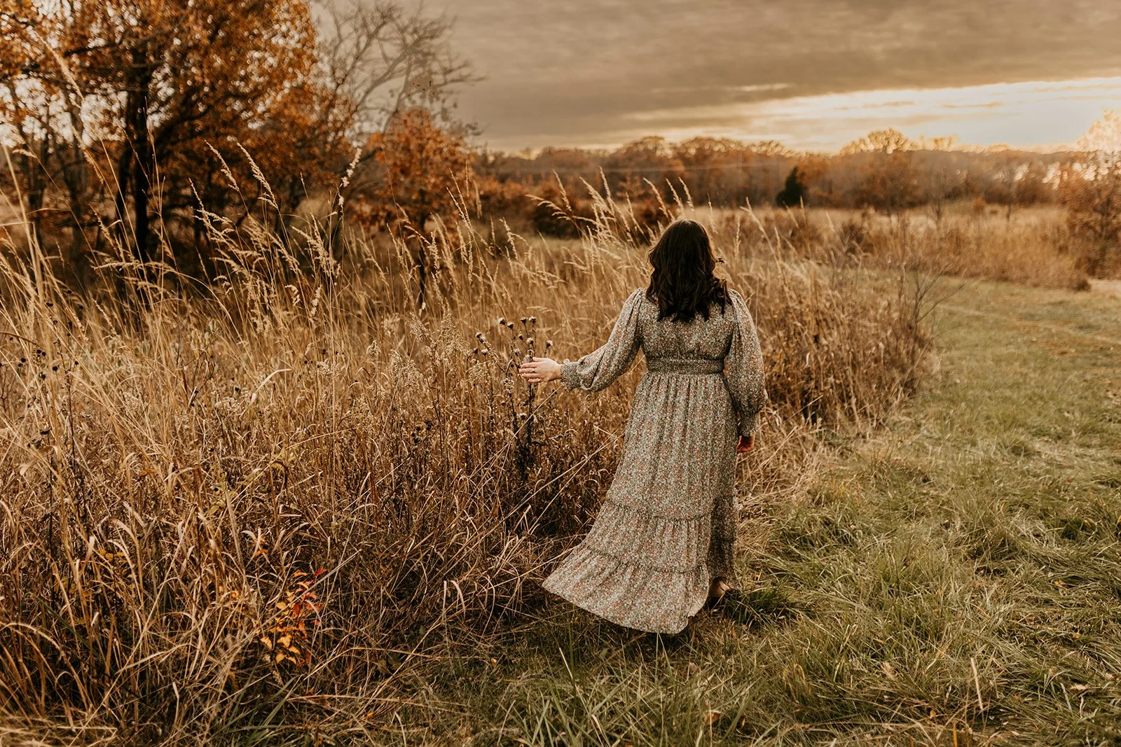 A woman with dark hair wearing a long floral dress walking on a grassy path through a field of tall dry grasses and trees with autumn-colored leaves, under a cloudy sky during sunset.