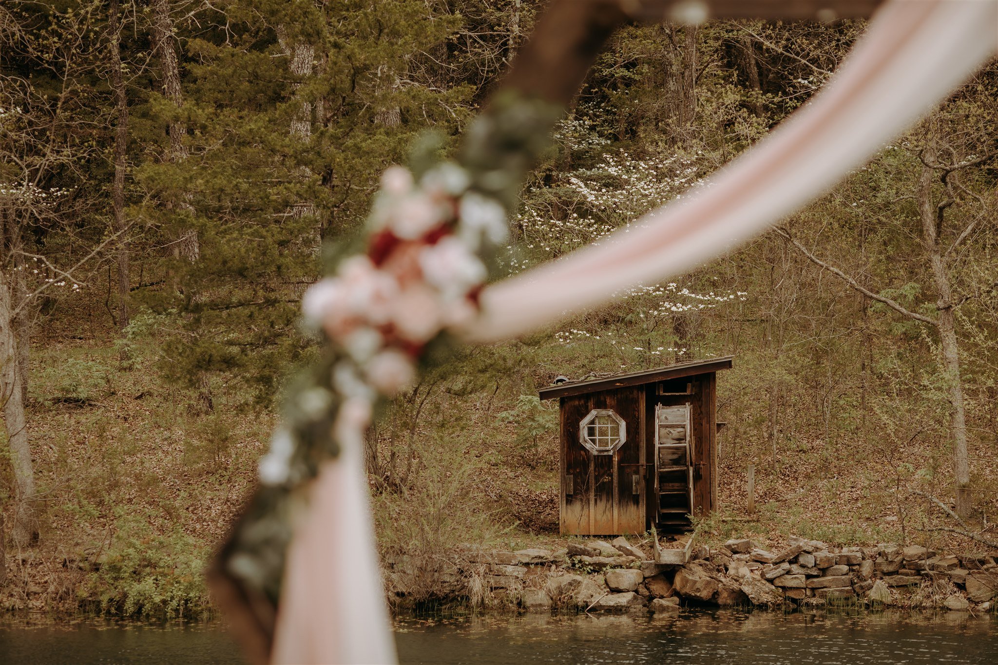 A small wooden cabin on a riverbank surrounded by trees, viewed through a blurred floral and fabric decoration, likely part of an outdoor event setup.